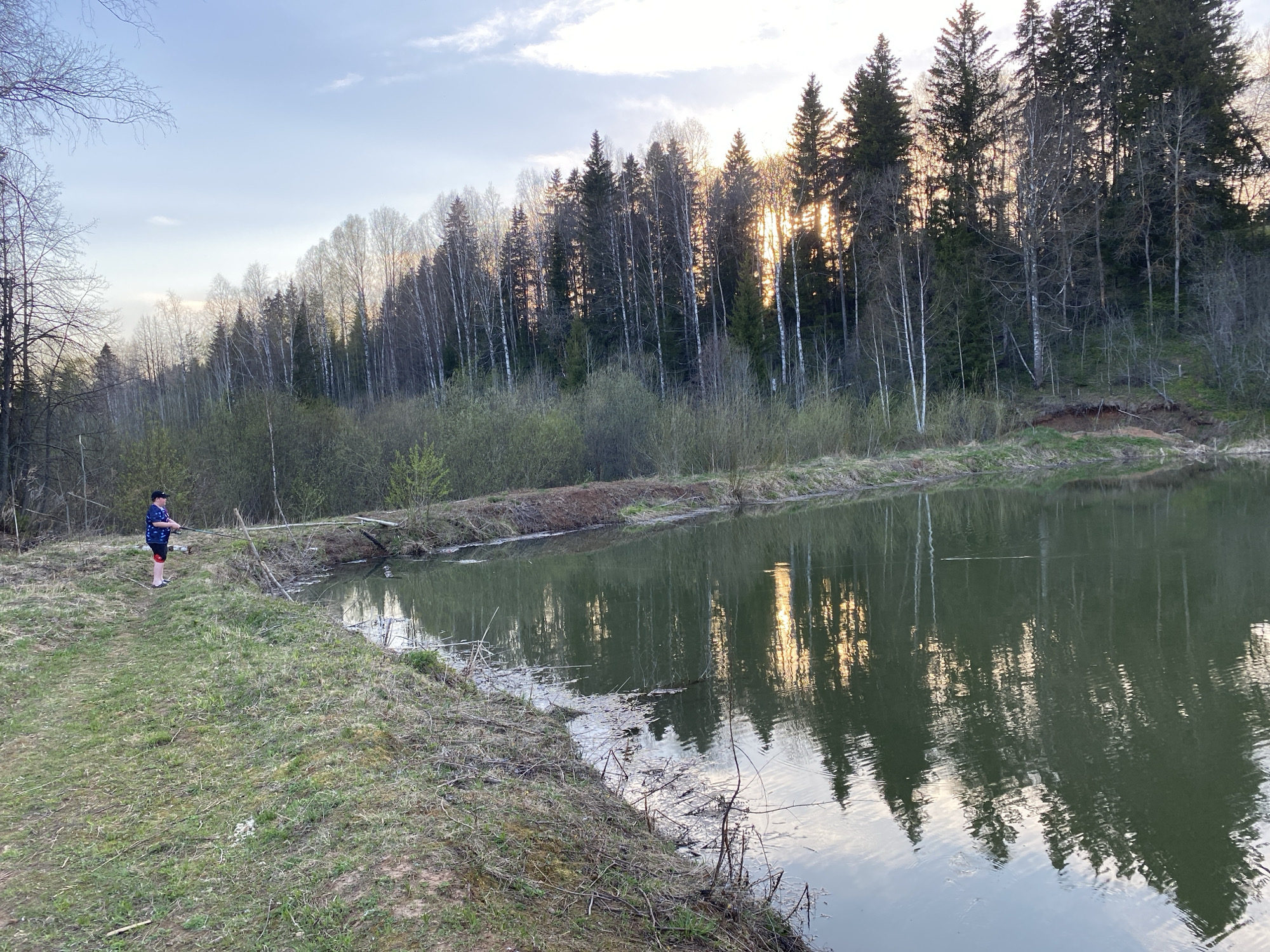 The image depicts a person fishing by a lake near Баклуши, Russia. The person, located on the left bank, wears a cap, a patterned blue shirt, and dark shorts. They hold a fishing rod, facing the water. The lake reflects the surrounding trees and sky. A forest, consisting of both deciduous and coniferous trees, lines the opposite bank. The sky is partly cloudy, suggesting either sunrise or sunset. The foreground shows grassy vegetation along the lake's edge.