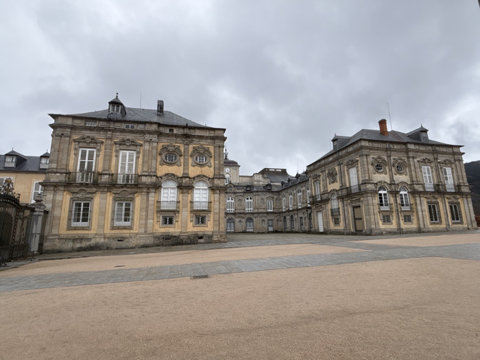 An expansive exterior view captures the Baroque architecture of the Royal Palace of La Granja de San Ildefonso in San Ildefonso, Spain, beneath an overcast, grey sky. The multi-winged palace features a primary facade constructed from light ochre-colored stucco with grey stone architectural details, including elaborate window frames, cornices, and quoins. Dark grey slate mansard roofs with dormer windows and chimneys cap the structures. Numerous windows, some with white louvered shutters, are visible; several on the first floor include small, ornate wrought-iron balconies. A clock tower is discernible in a recessed central section of the palace. The immediate foreground and midground present a vast, empty plaza surfaced with sandy-colored gravel, delineated by sections of grey stone paving. To the far left, an ornate metal gate is partially visible. The distant right background shows a dark, densely vegetated mountain slope. No individuals, actions, or interactions are present in the scene.