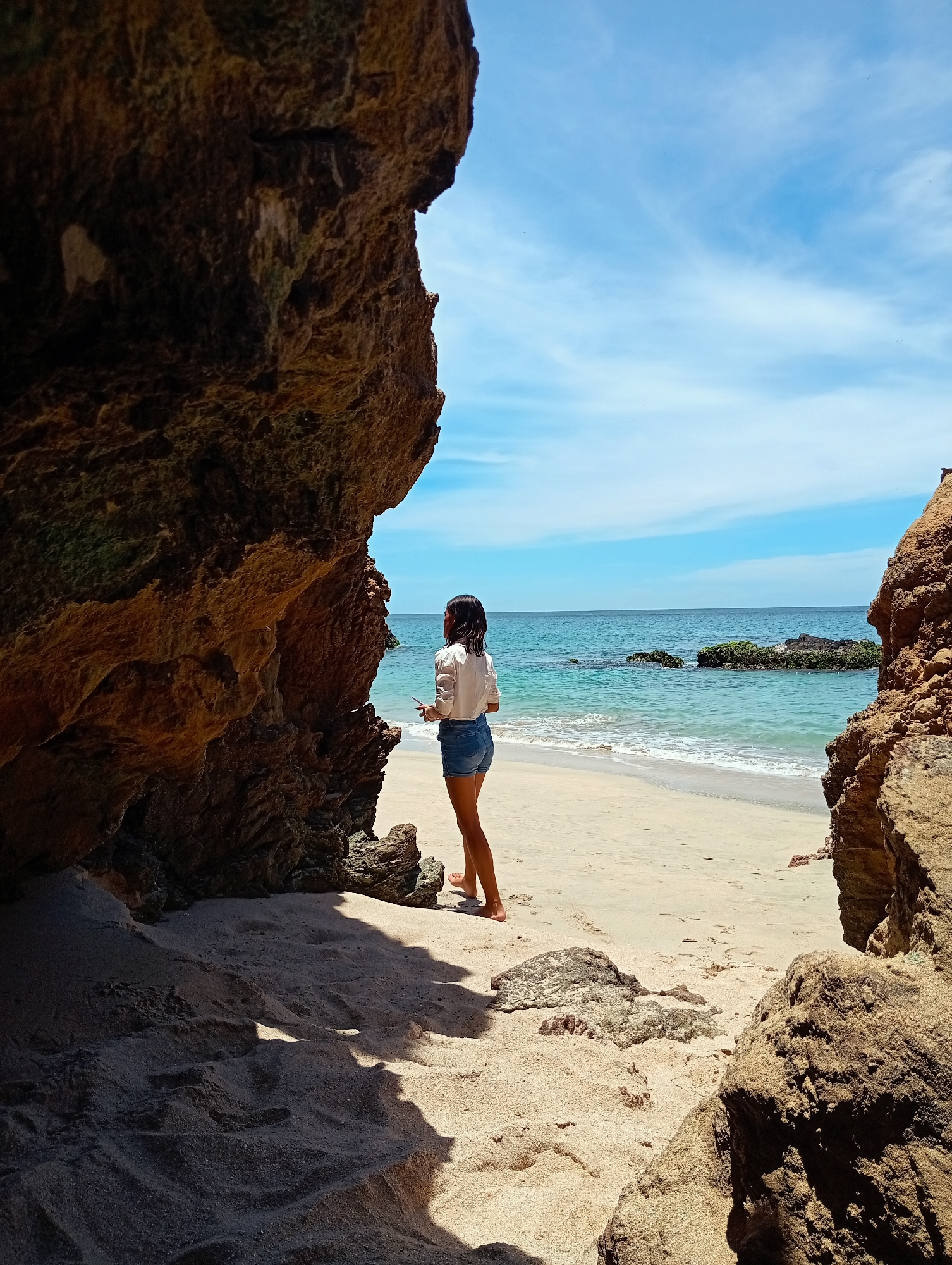 The image shows a woman standing on a beach at Playa Caribe in Pedro González, Venezuela. She is viewed from behind, wearing a white shirt and denim shorts. The woman is barefoot and appears to be looking out at the ocean. The scene is framed by two large rock formations on either side, which cast shadows on the sand. The ocean is visible in the background, with several rocky outcroppings extending into the water. The sky is blue with scattered clouds.