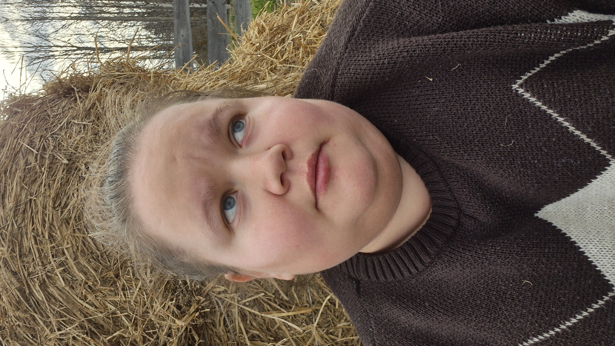 The image shows a close-up portrait of a person with light skin, blue eyes, and light brown or blonde hair, looking slightly upwards and to the left. They appear to be lying down, possibly on their back. Their expression is calm and neutral, with rosy cheeks. They are wearing a dark brown knitted sweater featuring a white argyle pattern. A few small strands of hay are visible on the sweater.
The person is situated against a large, golden-brown hay bale, which forms the immediate background. Beyond the hay bale, an outdoor scene unfolds under a bright, likely overcast sky. Bare tree branches are visible in the upper background, indicating a cooler season such as late autumn, winter, or early spring. A wooden fence post can also be seen in the distance, reinforcing the rural setting in Дойная, Russia. The soft, diffused lighting suggests it is daytime. No text is visible in the image.