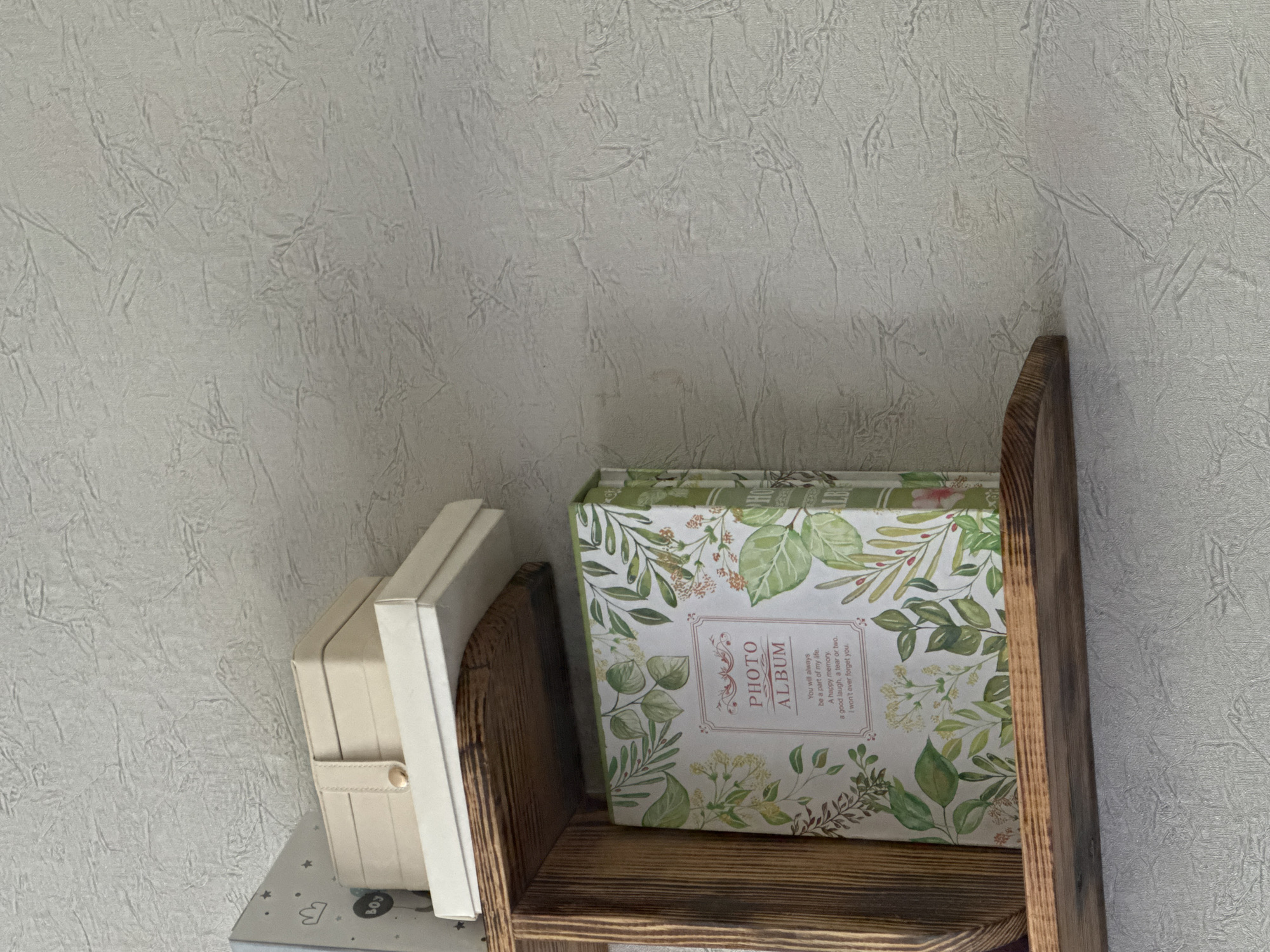 This image shows a close-up view of a wooden shelf containing two decorative boxes. The box on the right is a photo album with a floral pattern of green leaves and small white and yellow flowers. It has the text "PHOTO ALBUM" prominently displayed on its cover, along with a brief poetic inscription. The box on the left is a lighter-colored, possibly beige or cream, box with a strap and a small circular clasp. Both boxes are placed on a shelf with a textured, light-colored wall behind them. The lighting suggests an indoor setting, possibly in the daytime, with the light source coming from above and to the left. The scene appears to be a home environment, suggesting a personal collection or storage area. The image is cropped to focus on these items, with the wall and the edge of the shelf also visible. No people are present in the image.