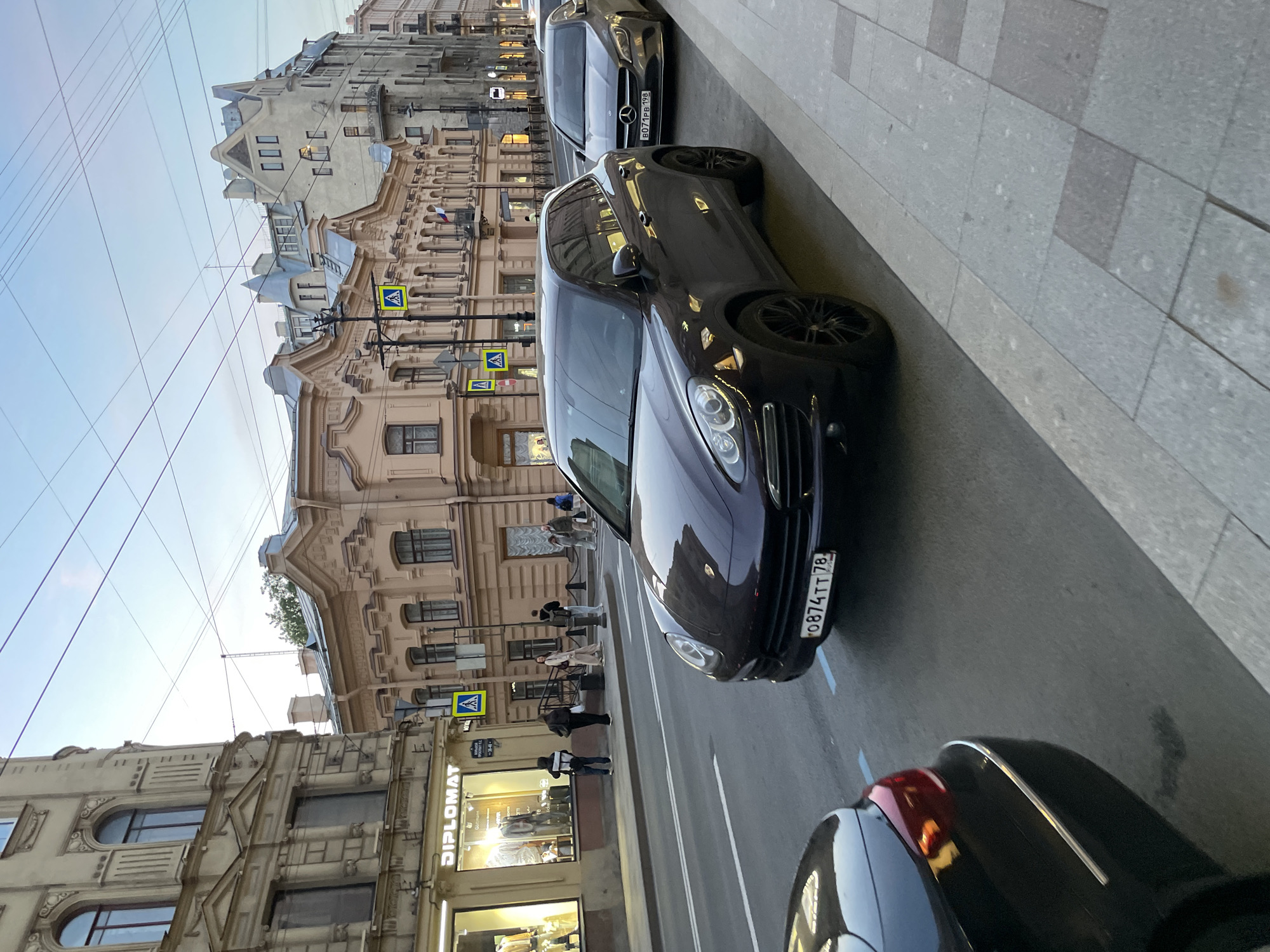 The image captures a street scene in Saint Petersburg, Russia. Several cars are parked along the right side of the road. The focus is on a dark-colored Porsche with a Russian license plate reading "O 874 TT 78". Another car, possibly a Mercedes-Benz, is parked ahead of it. The rear end of a dark car is also visible in the foreground.
Pedestrians are walking on the sidewalk, and some are standing near a store called "Diplomat." The buildings on the left side of the street feature classical architectural styles with decorative facades. Street signs indicating pedestrian crossings are mounted on poles. The sky is visible towards the top-left corner of the image, suggesting it's either morning or evening. Electrical wires run across the sky. The pavement on the right side of the road is made of large gray tiles.