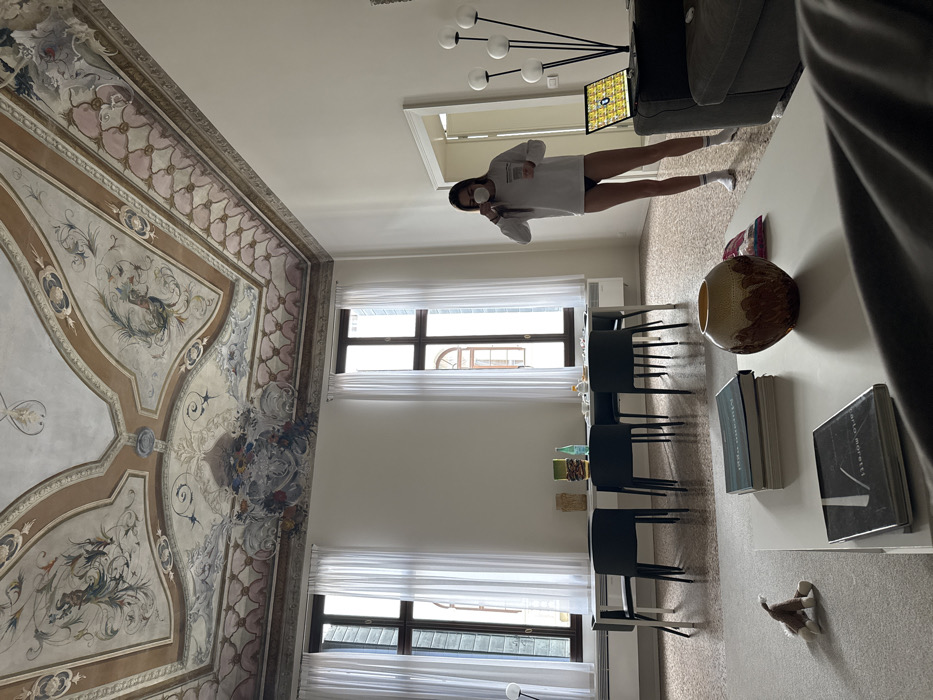 The image depicts a high-ceilinged room with an ornate frescoed ceiling, characteristic of a historic building in Venice, Italy. The ceiling features classical motifs, foliage, and avian designs in a muted palette of browns, golds, pinks, and blues, bordered by intricate patterns.
The room is furnished with a mix of contemporary and classic elements. Along the back wall, three tall windows, draped with sheer white curtains, offer views of adjacent Venetian architecture, including an arched window on an opposing building. Beneath these windows, a long, rectangular white dining table is positioned, surrounded by six dark, modern chairs. On the table, various items are visible, including a green bottle, a blue bottle, a yellow vase, and dinnerware. White radiator units are installed beneath the windows.
In the foreground, a white rectangular coffee table displays a stack of books, a textured brown and gold vase, and a small colorful item. To the left of the coffee table, on the terrazzo-style floor, a small stuffed animal is present. A dark gray sofa is partially visible at the bottom right.
A female individual stands on the right side of the room, wearing a long-sleeved white top, dark briefs, and white socks. She has long dark hair and a white face mask pulled down under her chin. She holds a white cup to her mouth, appearing to drink, while her right hand is near her ear, possibly holding a phone or earpiece. Adjacent to her, a modern floor lamp with multiple spherical white lights stands next to an open laptop displaying a colorful tiled pattern on its screen. An open doorway on the right wall leads to another room. The flooring throughout the room is a light-colored terrazzo with scattered darker aggregates.