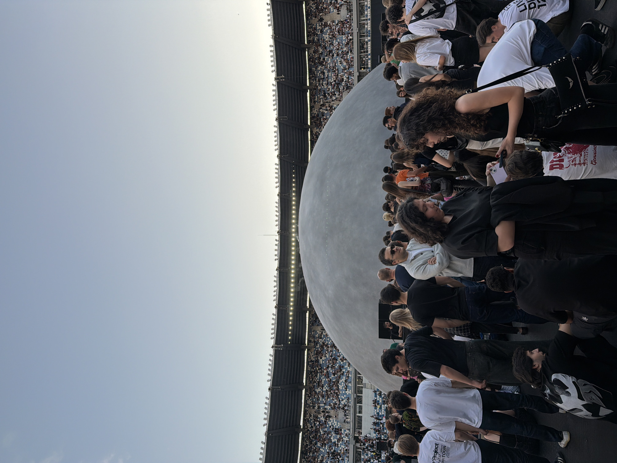 The image depicts a large outdoor gathering of people in front of a dome-shaped structure within a stadium. The stadium seating is visible in the background, filled with spectators. The sky above is clear and light blue. In the foreground, a crowd of individuals are standing and interacting. Some are looking at their phones, while others are engaged in conversation. The dome appears to be a central feature of the event. Based on the context of the structure and the stadium, this scene is likely taking place at the Mikheil Meskhi Stadium in Tbilisi, Georgia, which is known for hosting various events and concerts.