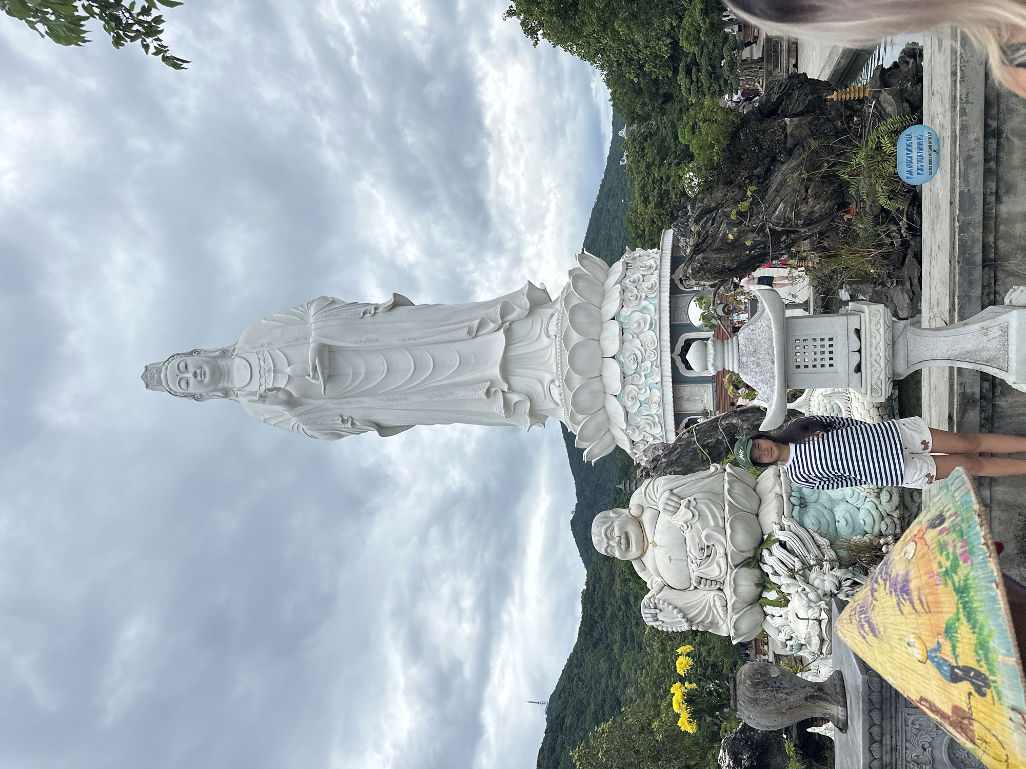 This image captures a view of the Linh Ung Pagoda in Đà Nẵng, Vietnam, featuring a prominent, large white statue of Quan Âm (Goddess of Mercy) standing on a lotus base. To the left of Quan Âm, a smaller, seated white statue of a laughing Buddha is visible. In the foreground, a young girl wearing a green cap, a striped long-sleeved shirt, and white shorts stands in front of a decorative conical hat. The environment includes lush green hills in the background, a cloudy sky, a stone lantern, and decorative rock formations near a body of water. A sign with Vietnamese and English text is placed on the ground near the rock formations, reading "QUAN KHÁCH KHÔNG NÊN ĐỨNG TRÊN THÀNH HỒ" and "VISITORS SHOULD NOT STAND ON THE G.TI".