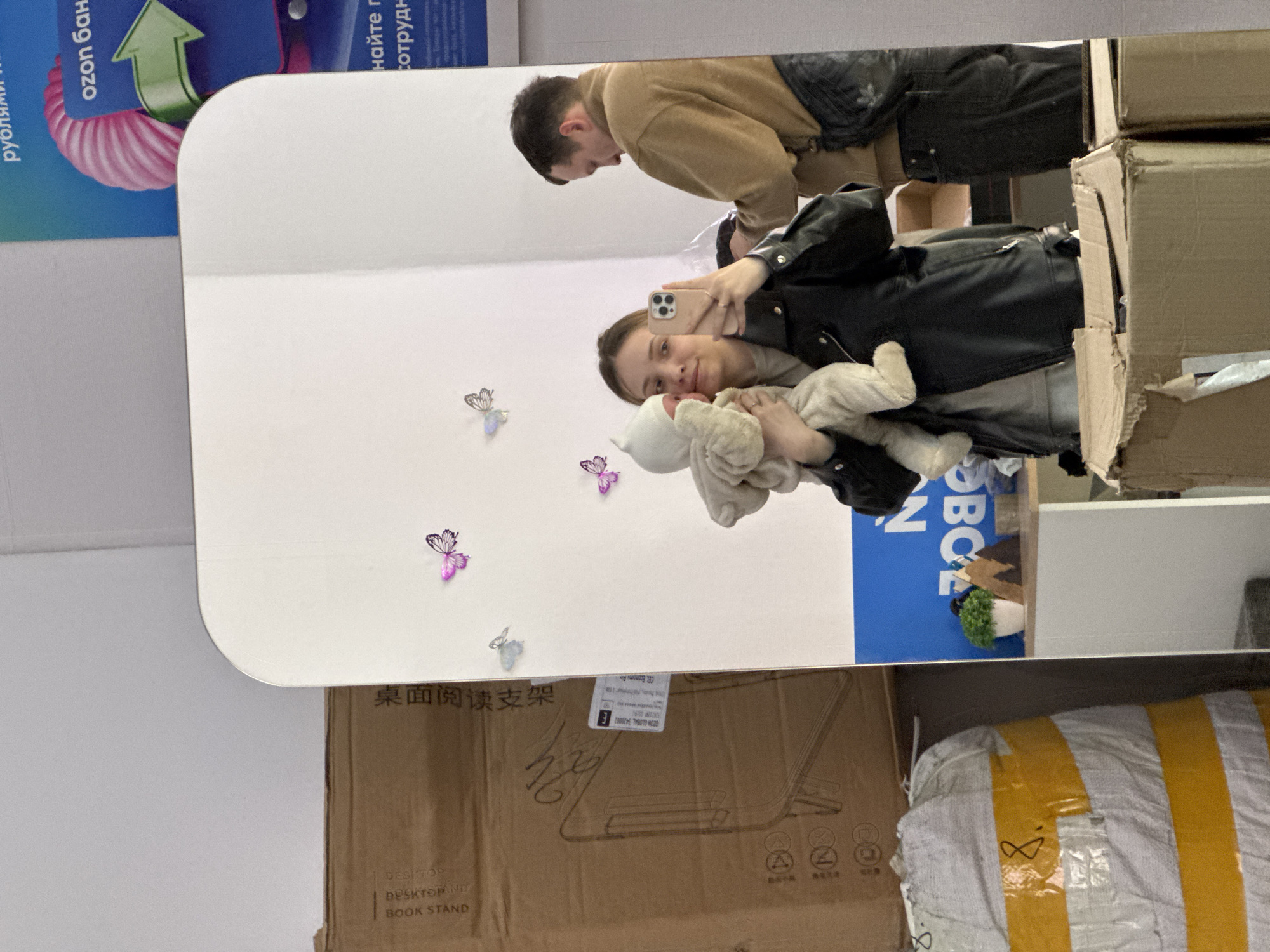 The image depicts a mirror reflection of a woman holding a baby, taking a selfie with her phone. In the background, a man is seen standing next to her. The mirror has decorative butterfly stickers on it. Below the mirror are various objects including cardboard boxes, one labeled "桌面阅读支架" (Desktop Reading Stand) and "BOOK STAND", and a large white and yellow striped package. The image also shows parts of an advertisement with text in Russian, "ozon бан" and "найте г сотрудн". The photograph appears to be taken indoors, possibly in a store or a storage area. There is no way to determine if this image was taken in the city of Chaikovsky, Russia.