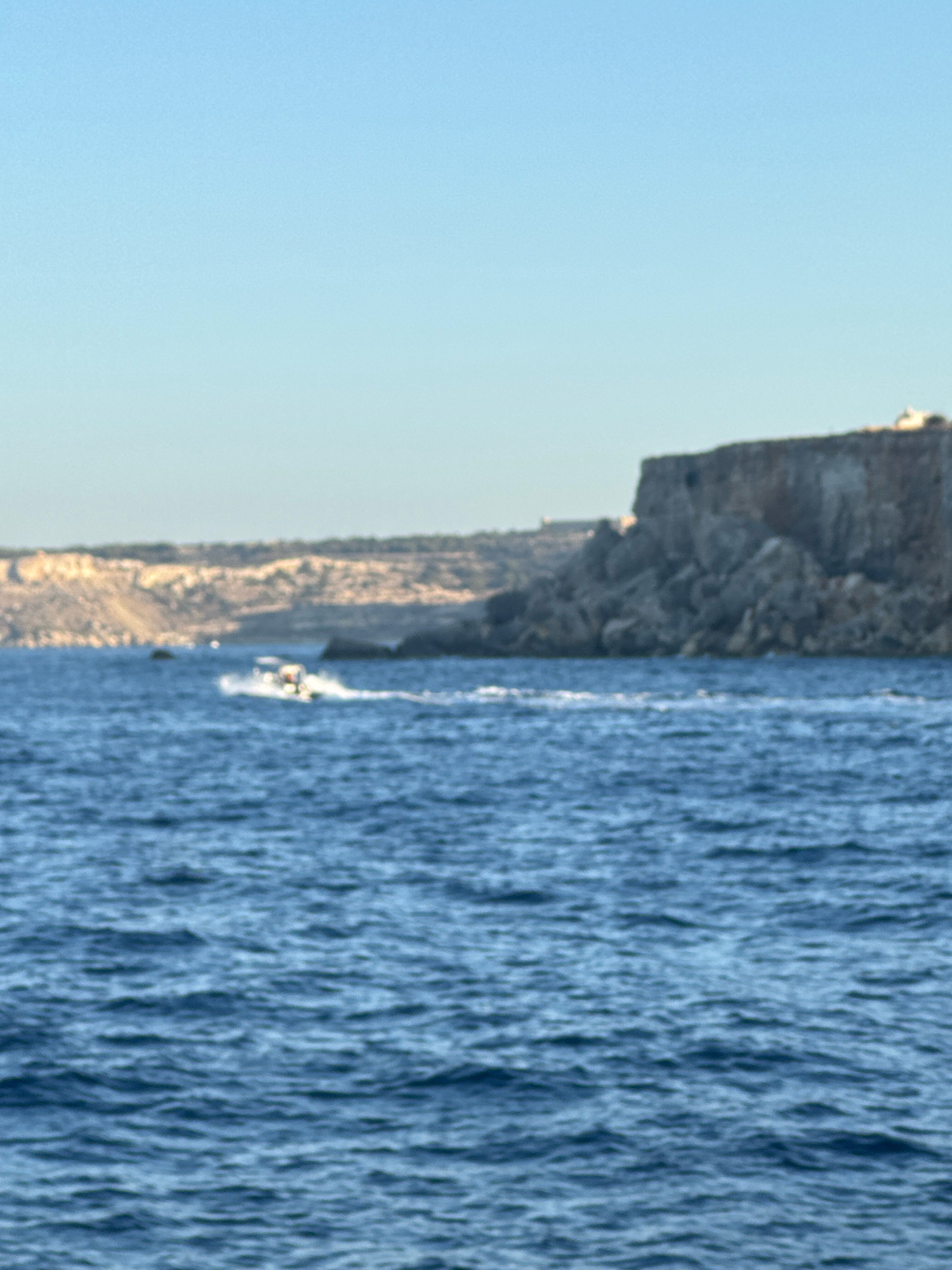 The image shows a boat speeding through the blue sea off the coast of Mellieħa, Malta. The boat is white with a yellow canopy and is creating a white wake behind it. In the background, a rocky cliff face rises from the sea, with some vegetation visible on top. The sky is clear and blue. The image appears to have been taken from another boat.