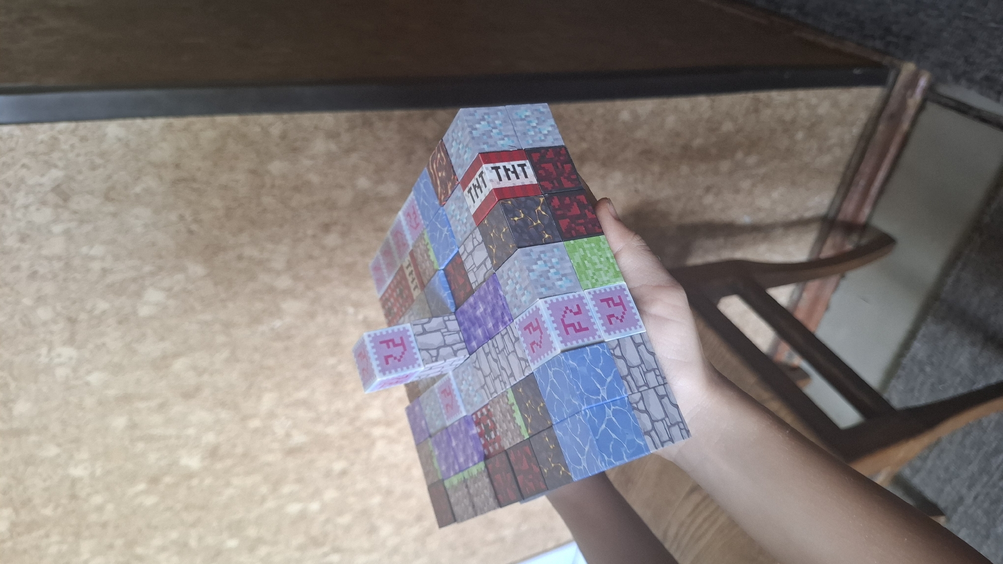 A close-up, high-angle shot shows a multi-layered structure of Minecraft-themed blocks, held by a person's hands in the foreground. The blocks feature various textures and colors, including stone, TNT, and what appear to be different types of ore. The background is a textured wall, likely indoors, with a portion of a dark vertical frame visible on the right. No specific location in Shymkent, Kazakhstan, can be identified.