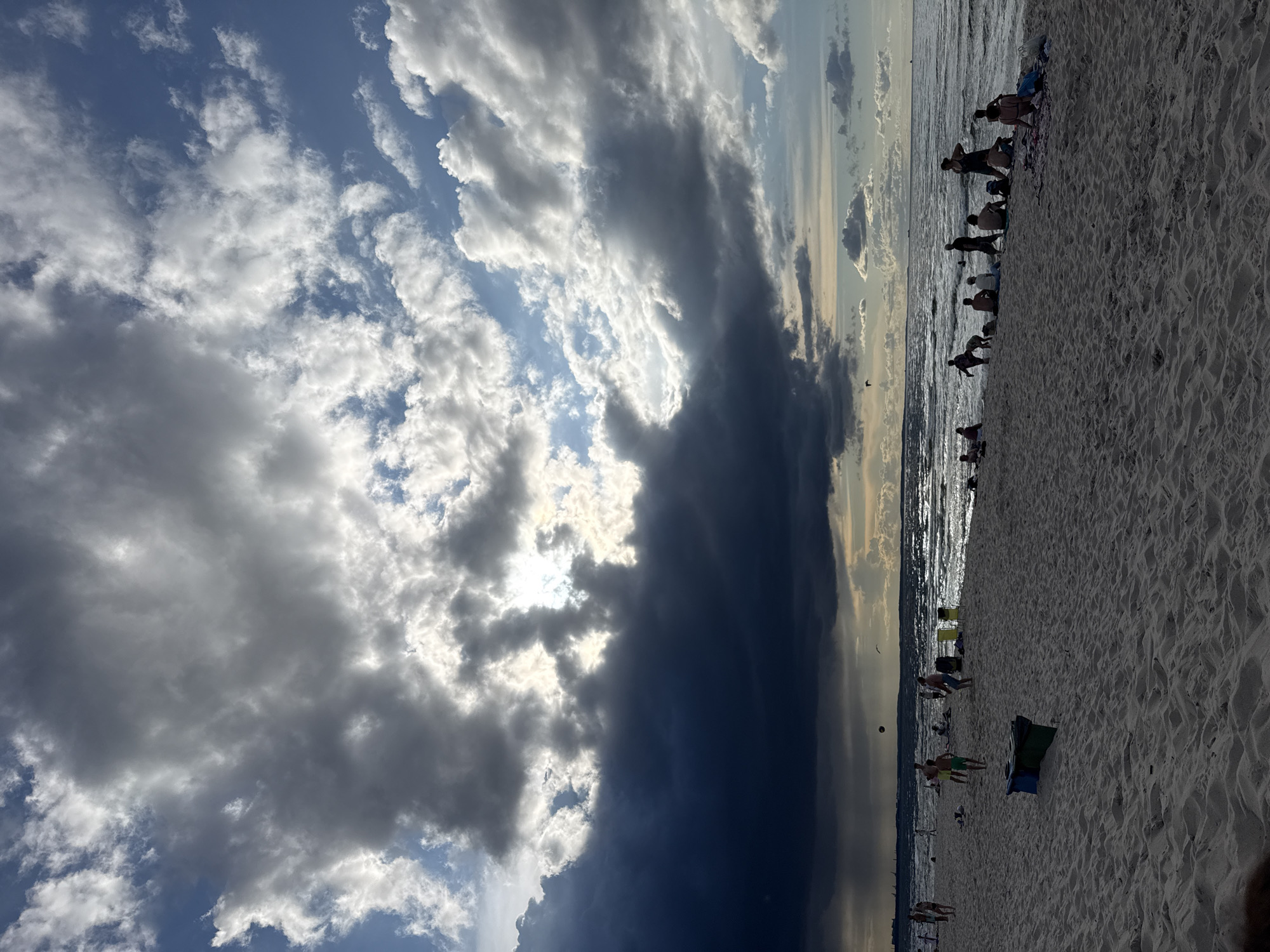 The image depicts a sandy beach in Międzyzdroje, Poland, under a dramatic sky filled with both dark, stormy clouds and areas of bright sunlight. A body of water, likely the Baltic Sea, stretches to the horizon, with waves breaking on the shore. Several individuals are visible on the beach, some are sitting or lying on towels, while others are standing or walking. A group of people appears to be engaged in a game of volleyball in the middle ground. In the foreground, there are two trash receptacles, one green and one blue. The lighting suggests it is late afternoon or early evening, with the sun intermittently breaking through the heavy cloud cover.
