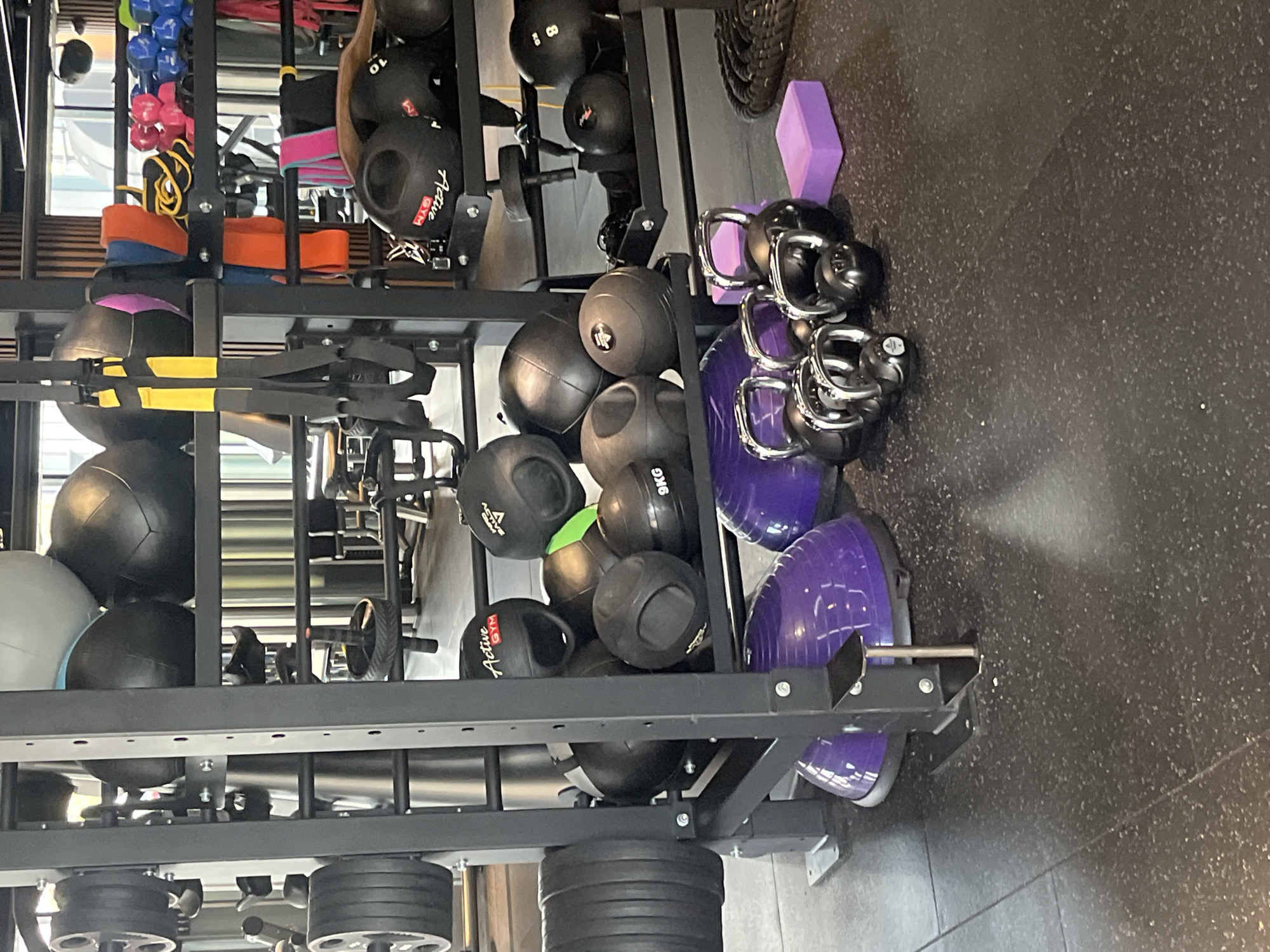 This image captures a dedicated storage area within an indoor gym located in Sofia, Bulgaria. The central focus is a multi-tiered black metal rack, systematically organizing a variety of fitness equipment.
On the upper shelves, sets of colorful dumbbells in blue and pink are visible, alongside resistance bands in vibrant orange, yellow, and purple hues. Lower sections of the rack and surrounding floor areas are densely packed with different types of balls: numerous black medicine balls or slam balls, some bearing the branding "Active GYM" and weight markings like "9KG" and "8". A grey stability ball is also partially visible. A cluster of black kettlebells, some with shiny silver handles and one marked "40LBS", are stored near purple stability balls, which are also neatly arranged on the floor. A light purple yoga block rests on the dark, speckled gym flooring next to the kettlebells. Other equipment includes an ab roller, TRX suspension trainers, and black weight plates stacked on the lower part of the rack.
The space appears well-lit, suggesting daytime or bright artificial lighting. The equipment is neatly organized, indicating a well-maintained and functional gym environment. No people are present, and thus no specific activity is taking place, but the array of gear is ready for various workouts.