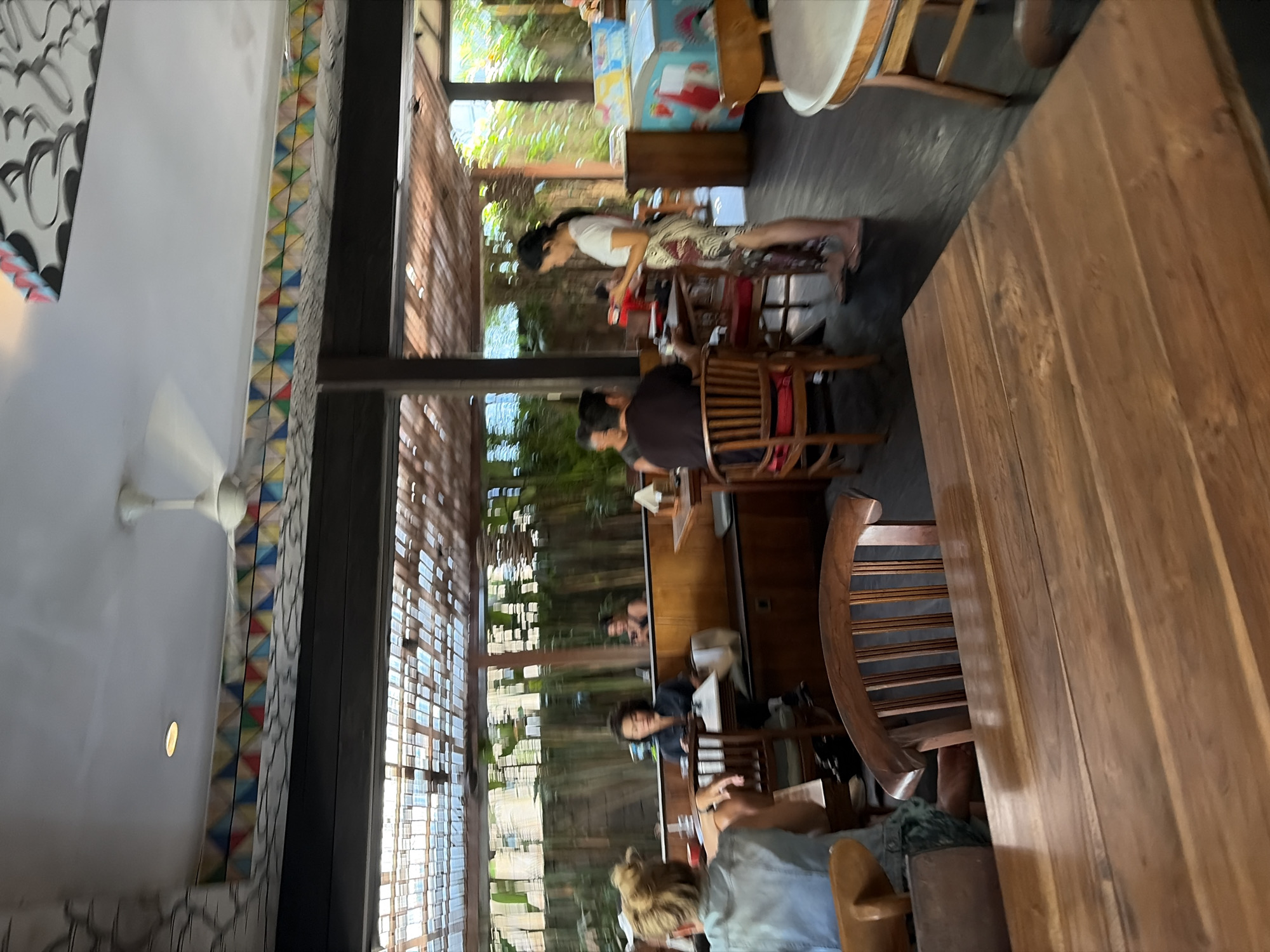 This image depicts the interior of an open-air restaurant in North Kuta, Indonesia. The scene is characterized by its rustic and natural ambiance, with wooden furniture and tropical foliage visible in the background. Several patrons are seated at tables, engaged in conversations and dining. A woman in a white top and patterned skirt stands near a refrigerator decorated with images of fruit, possibly serving or attending to customers. The ceiling features a decorative border with colorful geometric patterns and a ceiling fan. Natural light filters through the slatted wooden roof, illuminating the dining area.