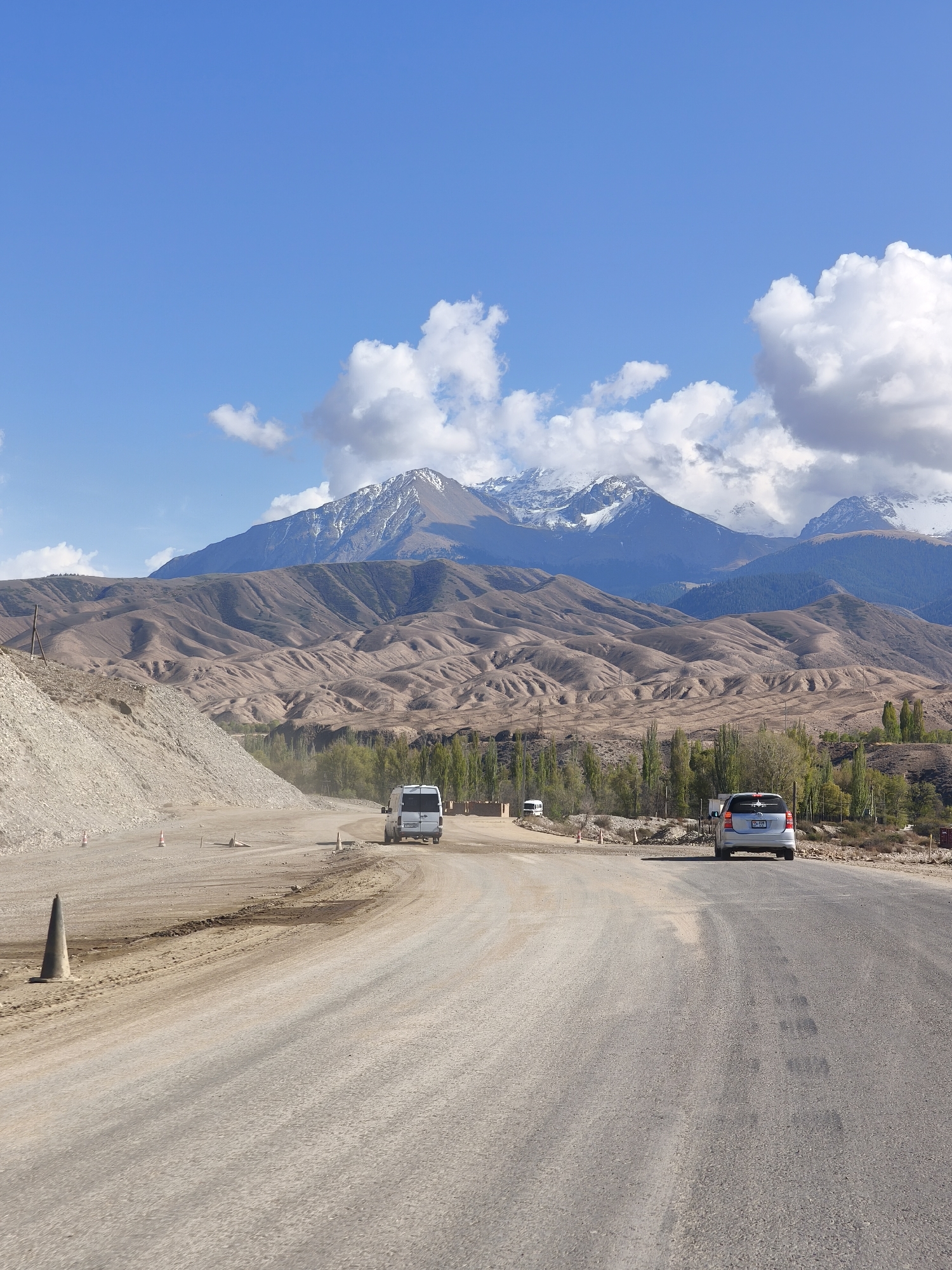 The image presents a vast, rugged landscape near Tamga, Kyrgyzstan, under a bright blue sky dotted with scattered white clouds. A wide, partially paved and partially dirt road dominates the foreground, curving gently to the right. The road surface appears dusty and uneven in sections. To the left of the road, a large pile of light-colored earth and rocks suggests ongoing construction or earthwork. Several orange traffic cones are visible along the roadside, indicating a temporary lane or work zone.
Two vehicles are traveling away from the viewer on the road. In the distance, a white panel van moves along a dirt section of the road. Closer to the foreground, a silver compact SUV or minivan is visible on the paved portion. Its rear license plate clearly displays "09KG 927 AEL," with "KG" indicating Kyrgyzstan.
The midground features rolling, barren hills, sculpted into soft, undulating forms, stretching across the scene in shades of brown and beige. A line of tall, slender green trees, likely poplars, grows between the road and these hills. The background is dominated by magnificent, snow-capped mountains, their peaks bright against the sky, indicating a high-altitude environment. The scene depicts a sunny day, suggesting fair weather for travel through this impressive natural setting.