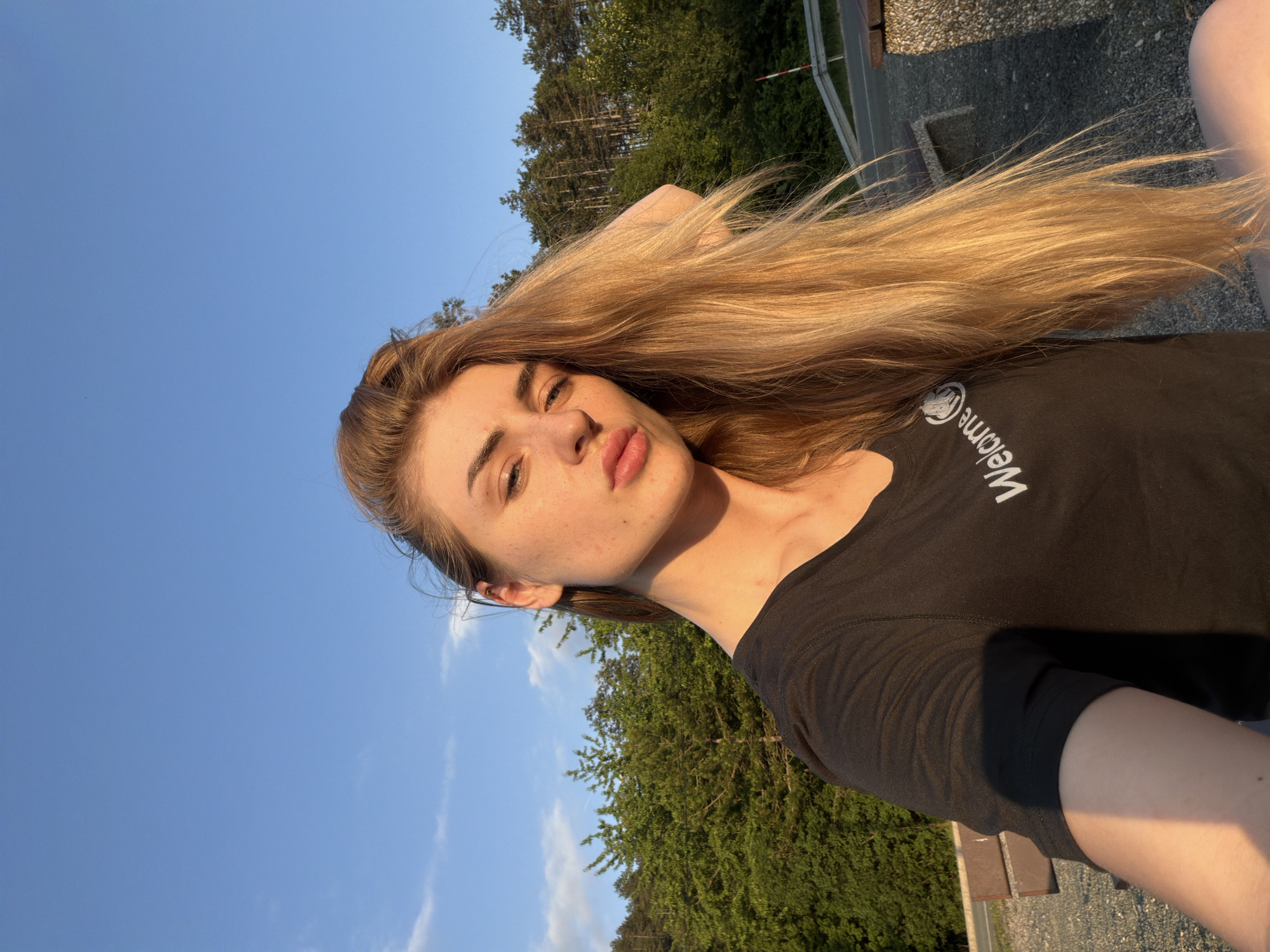 A young Caucasian woman with long, blonde hair is pictured outdoors in a selfie. She is wearing a black t-shirt with the word "Welcome" printed on it in white. Her hair is flowing and catches the sunlight, highlighting its golden tones. She has freckles across her nose and cheeks. The background consists of lush green trees and a bright blue sky with a few wispy clouds. A road and a bench are partially visible in the distance, suggesting an outdoor recreational area or a scenic overlook. The lighting indicates it is late afternoon or early evening, with the sun casting a warm glow on the scene.