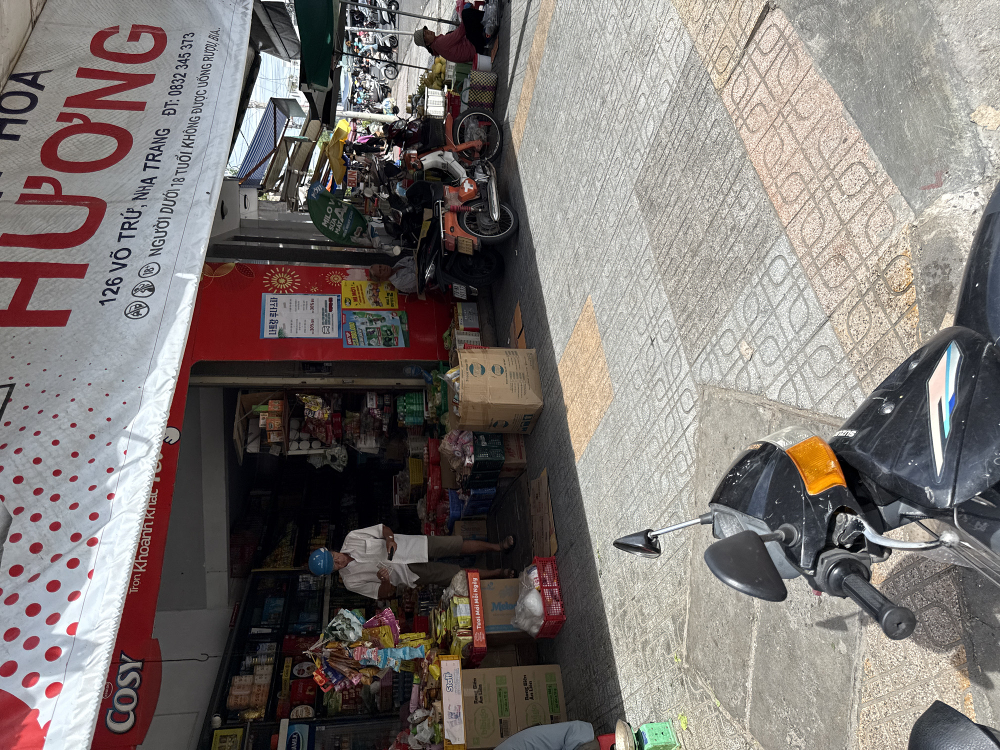 In front of a shop in Nha Trang, Vietnam, a man wearing a white shirt and blue hard hat stands looking at his phone. The shop entrance displays a red and white awning with red polka dots and a large red sign with white lettering that reads "HƯƠNG HOA". Below this, smaller text indicates the address "126 VÕ TRỪ, NHA TRANG" and a phone number. Shelves inside the shop are stocked with various goods. To the right of the shop entrance, an orange and white motorcycle is parked. Further to the right, a woman in a pink shirt and straw hat is seated on the ground near a stall displaying fruit. The ground is paved with patterned tiles. A black motorcycle is partially visible in the foreground of the image.