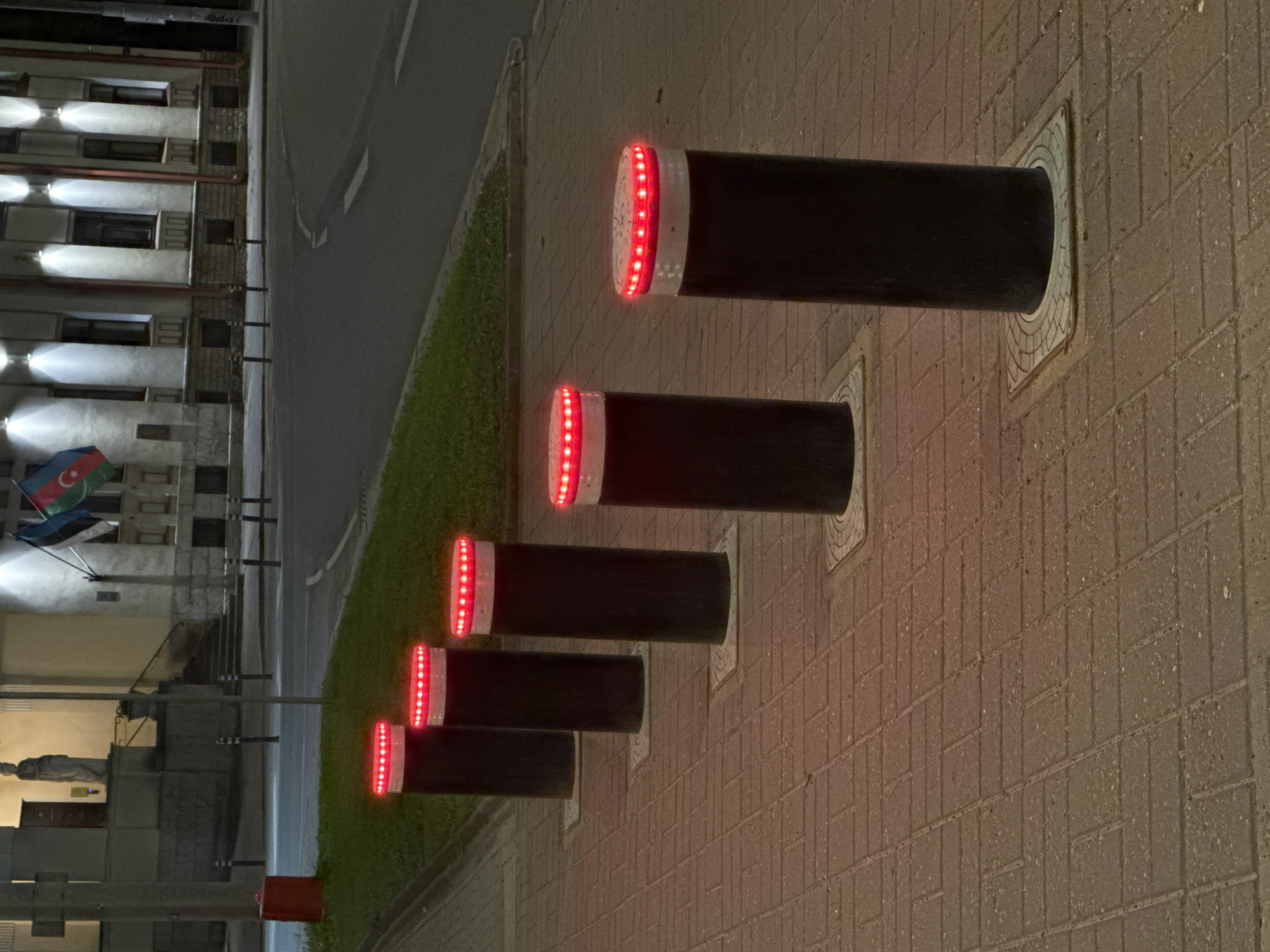 A street scene at night in Tallinn, Estonia features a paved sidewalk with several black cylindrical bollards spaced evenly apart. The bollards have a white band near the top, with a ring of red LED lights illuminating the top edge. In the background, a building with illuminated windows and architectural details stands behind a strip of grass and a street. Flags, including the flag of Estonia, are visible on poles in front of the building. A red fire hydrant is also present on the sidewalk. The road in front of the building shows traffic markings.