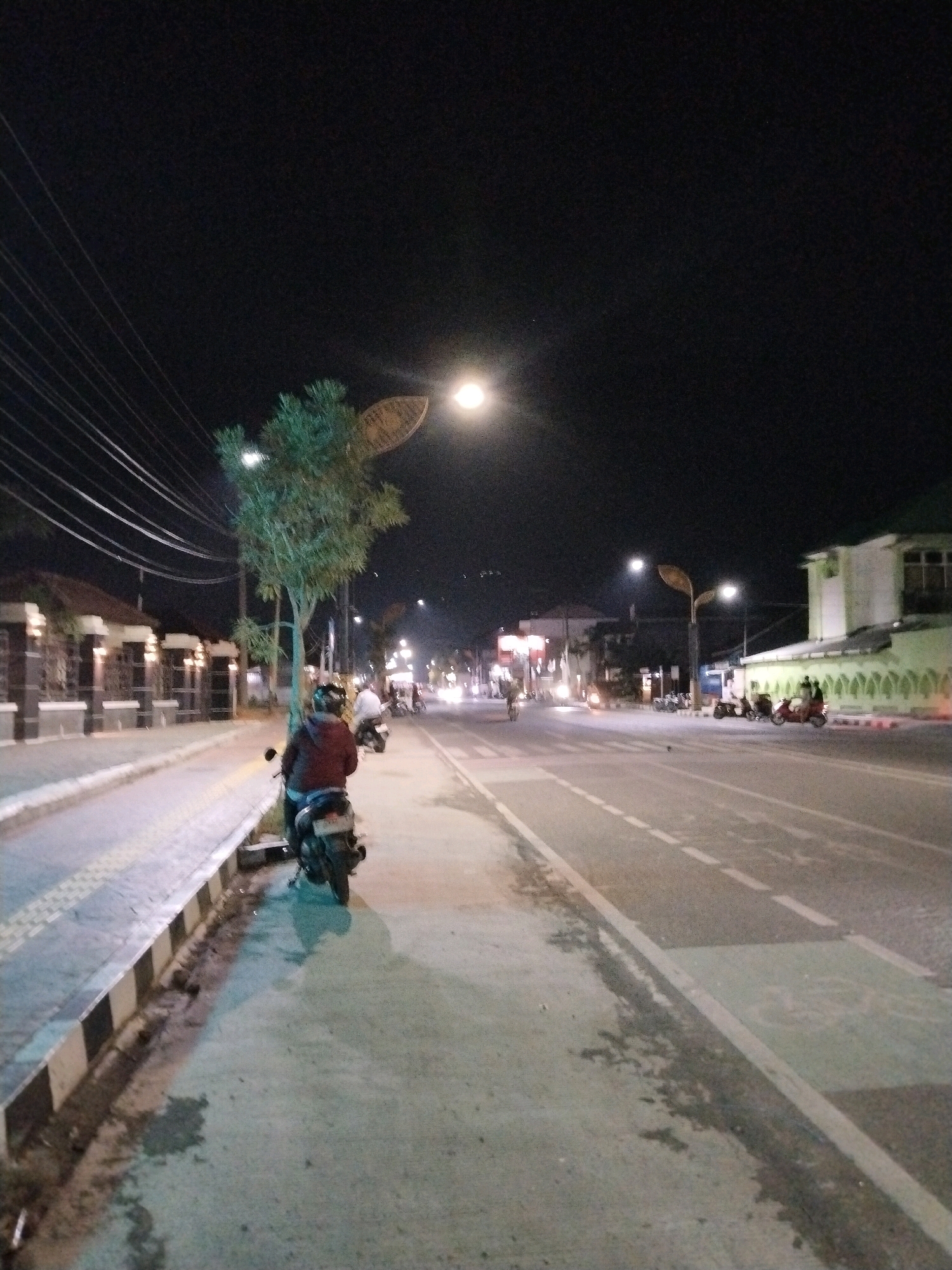 The image captures a street scene at night in Slawi, Indonesia. In the foreground, a person on a scooter is parked on a sidewalk. The sidewalk is paved with checkered tactile paving and has a curb with black and white stripes. A street lamp illuminates the area. Further down the sidewalk, a tree with a distinctive leaf-shaped lampshade is visible. Across the street, there is a road with traffic lanes marked by white lines. Several motorcycles are parked on the side of the road, and a building with a light green facade and decorative patterns is visible. The sky is dark, indicating nighttime. The overall scene depicts a typical streetscape in an Indonesian city at night, with elements of urban infrastructure and activity.
