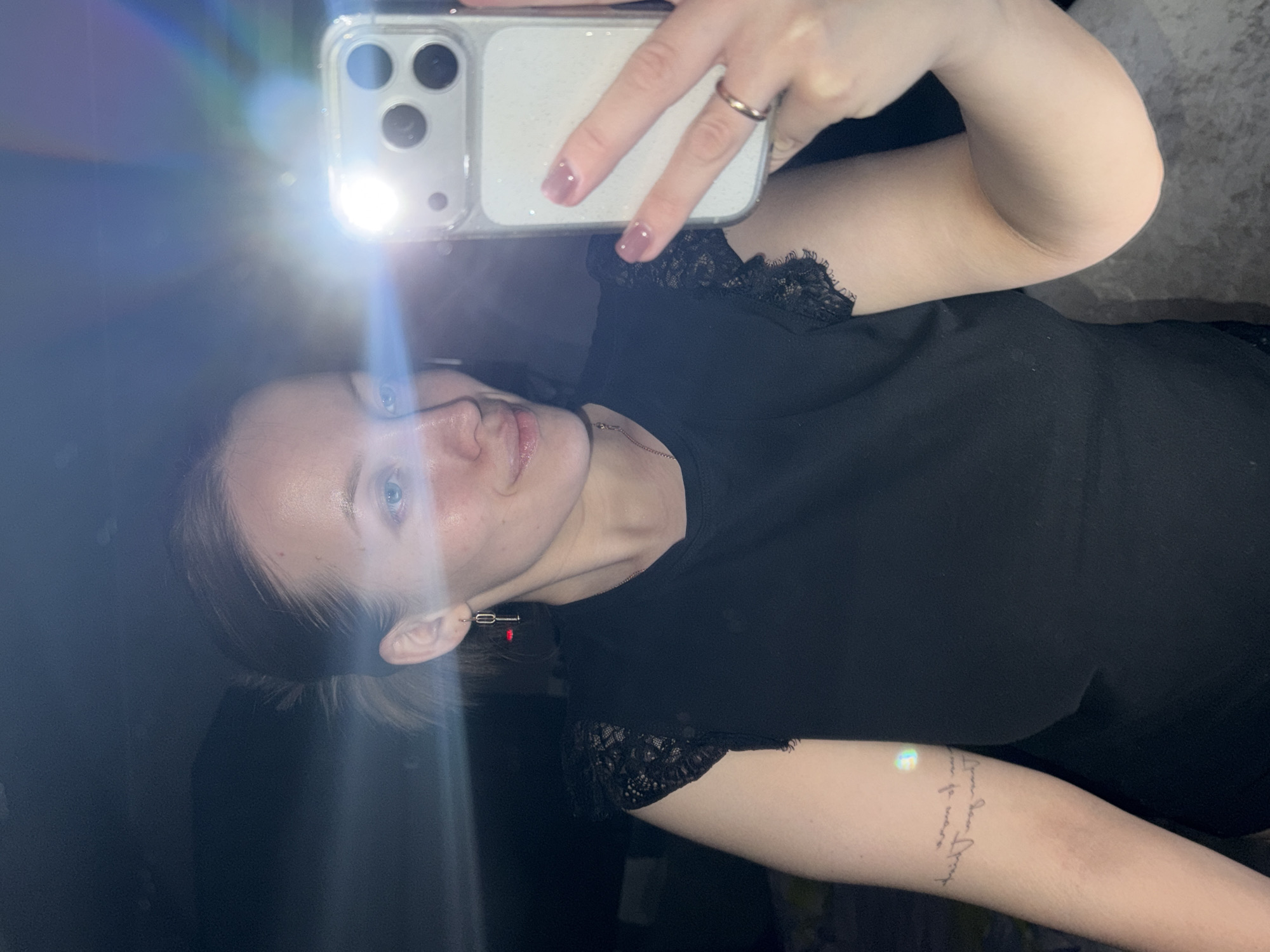A selfie taken by a light-skinned woman with blue eyes and light brown hair. She is holding a white iPhone in her right hand. The phone's flash is on, creating a bright glare in the image. The woman is wearing a black top with lace detailing on the sleeves. A silver ring is visible on her right ring finger. An earring is seen on her left ear. A tattoo with writing is visible on her right forearm. The background appears dark and indistinct. There is no evidence in the image to place this in Reshetnikovo, Russia.