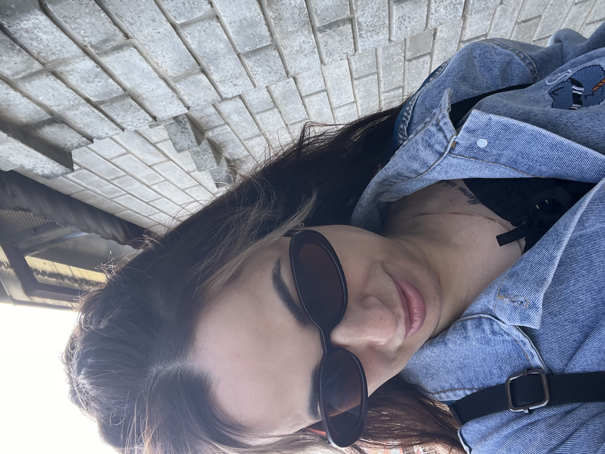 A woman wearing sunglasses and a denim jacket is taking a selfie outdoors, with a brick wall in the background. The woman has long, dark hair with some lighter streaks. She is looking down and smiling slightly. The denim jacket is blue and appears to have an embroidered patch on the front. The brick wall is light gray and appears to be a modern construction. The sky is visible in the upper left corner, suggesting it is daytime.