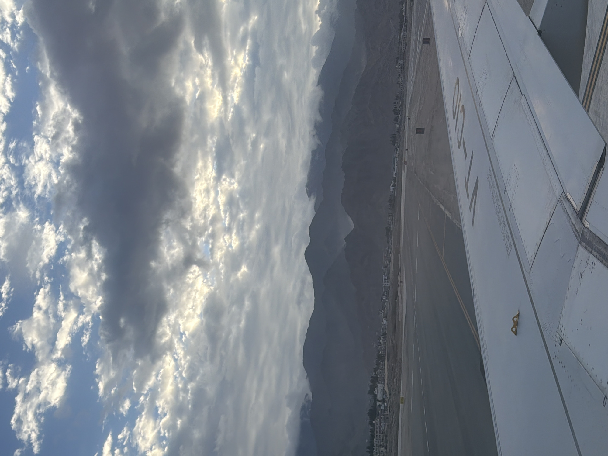 An airplane wing is in the foreground, partially obscuring a view of a tarmac and a distant mountain range under a cloudy sky. The wing is light gray and shows some rivets and markings, including the registration VT-CIO in yellow. The tarmac is a darker gray with yellow and white lines. In the middle ground, a town with buildings is visible nestled at the base of the mountains. The mountains themselves are dark and appear hazy, with some clouds lingering around their peaks. The sky is a mix of blue and gray clouds, with sunlight breaking through in some areas, suggesting either dawn or dusk. The scene appears to be at an airport near Spituk, India, given the presence of mountains characteristic of the region and the visible tarmac infrastructure. There are no people or active actions depicted in the image.