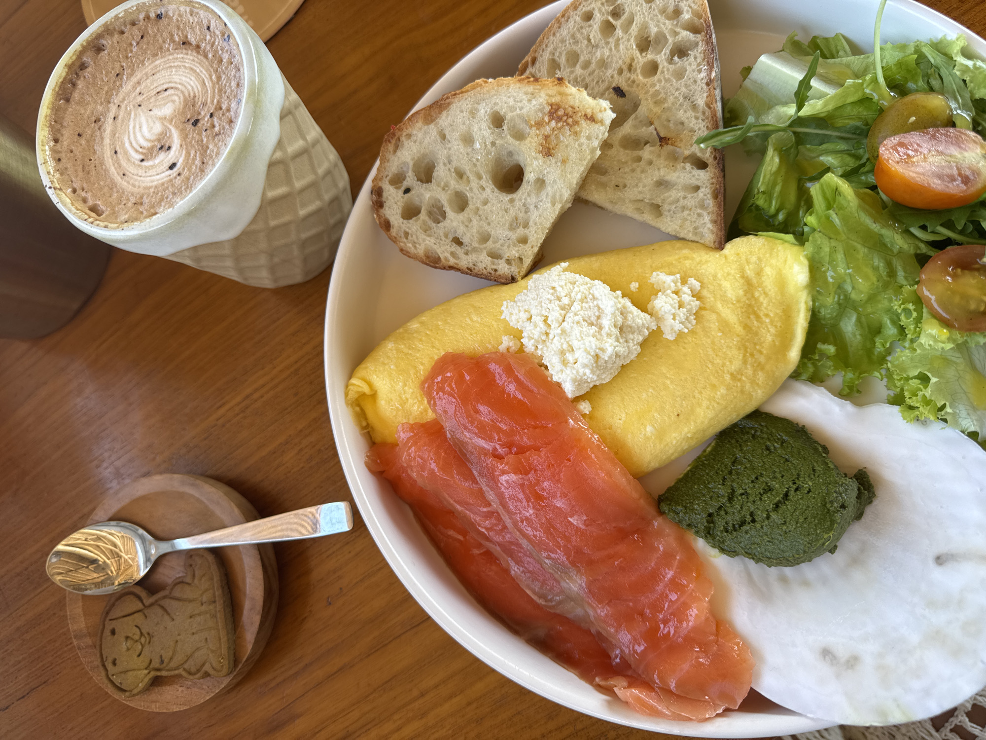 A high-angle shot presents a breakfast or brunch composition on a wooden surface. A white plate holds an assortment of food items: sliced bread, a rolled omelet topped with a dollop of cheese, slices of salmon, a green spread of pesto, a white radish slice, and a small salad with cherry tomatoes. Adjacent to the plate, there's a mug filled with a latte, displaying latte art on top. To the left, a wooden coaster holds a spoon and a small cookie shaped like a cartoon character. The setting suggests a cafe or restaurant, possibly located in an unknown city and region.