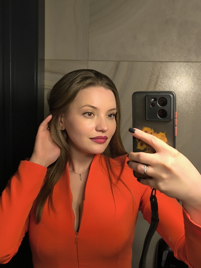 A woman in an orange long-sleeved dress takes a selfie in a bathroom. She holds a phone with a sunflower design on the case. Her hair is styled loosely around her face, and she wears lipstick. A silver cross necklace is visible. Behind her, a dark panel and light-colored tiled walls are seen.