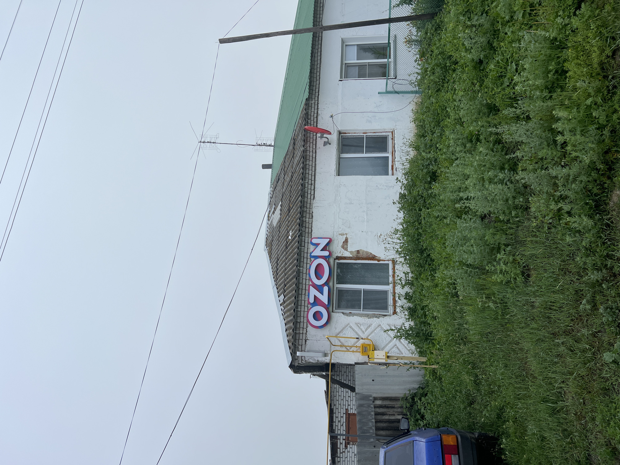 The image shows the exterior of a small, white, single-story building with a corrugated roof, appearing to be a retail establishment. A large, illuminated sign reading "OZON" in prominent blue and pink letters is affixed to the front of the building above a window. The building has two visible windows on its front facade, both with white frames. A green satellite dish is mounted on the wall between the windows, and a television antenna is visible on the roof. A yellow gas pipe runs along the exterior wall. The building is surrounded by overgrown green vegetation, and a blue car is partially visible on the left side of the frame. A wire fence runs along the right side of the property. The sky is overcast and grey, suggesting cloudy weather. The scene is located in Pivkino, Russia.