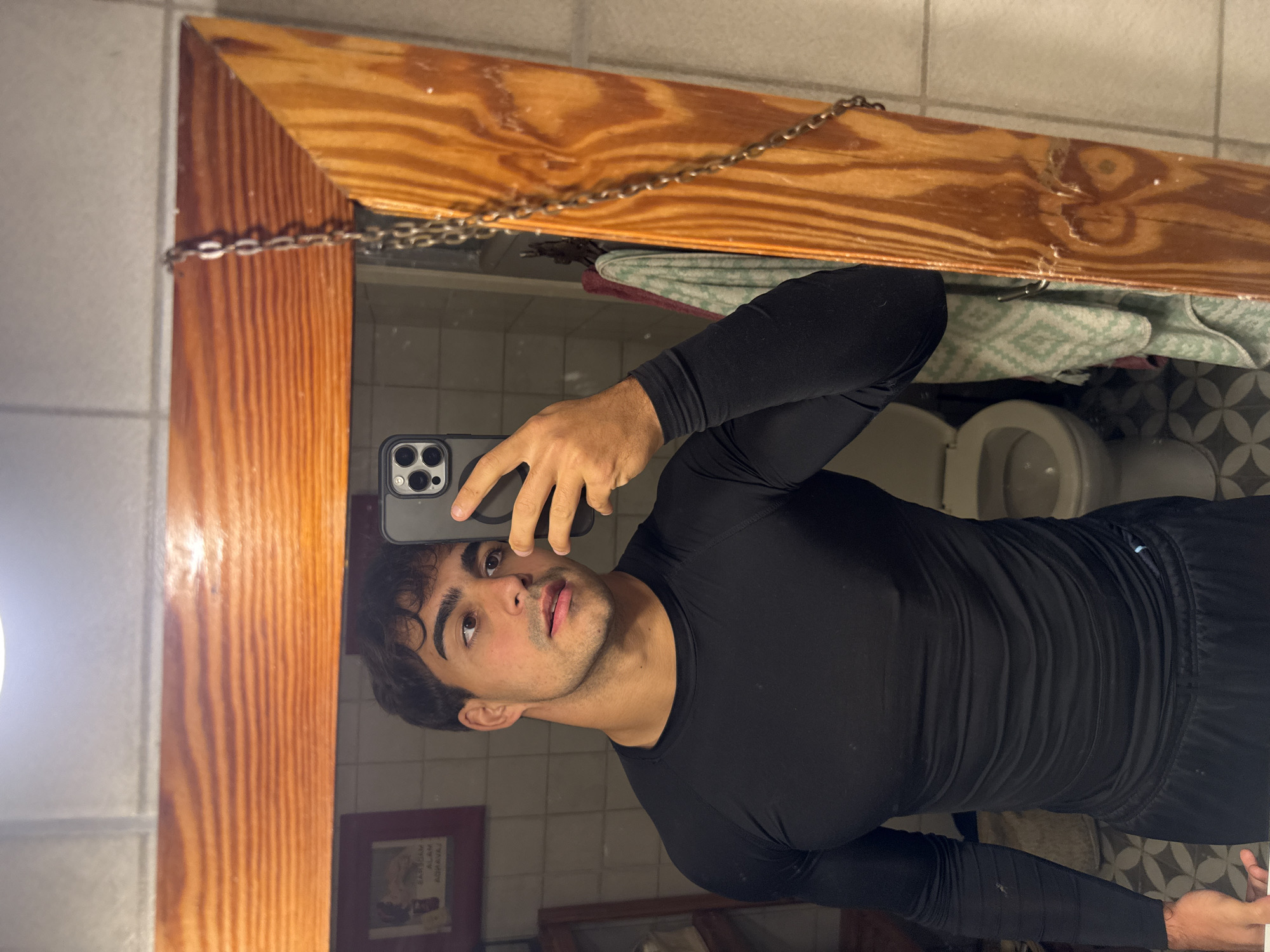 A young man with dark hair and brown eyes is taking a mirror selfie in a bathroom. He is wearing a black long-sleeved shirt and black shorts. The bathroom appears to have white tiled walls and a wooden-framed mirror. A toilet is visible in the background, as are a few towels hanging on a rack. The man is holding a smartphone in his right hand, which is reflected in the mirror. The scene is well-lit, with light coming from above. The image appears to be taken in a residential setting. This specific location in Avellaneda, Argentina, is a bathroom.