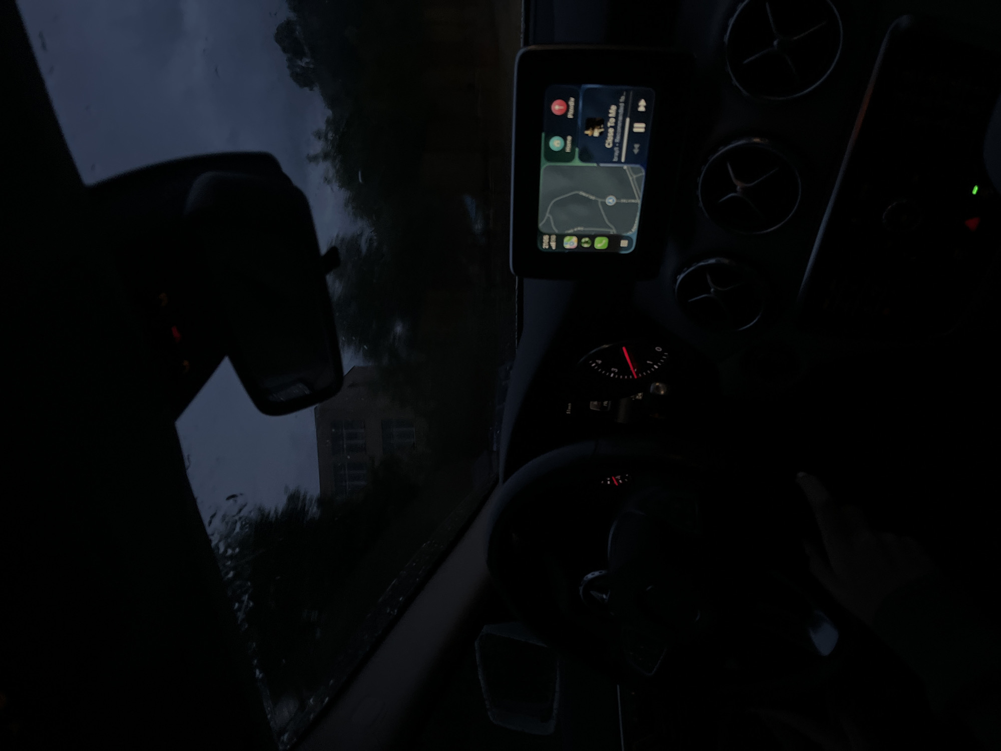The image depicts the interior of a car, likely a Mercedes-Benz, at night in Blagoevgrad, Bulgaria. Rain is visible on the windshield, and a building is discernible through the wet glass. The car's infotainment system displays a map with a route, likely for navigation. The speedometer needle is slightly above zero, suggesting the car is stationary or moving very slowly. The gear selector is in the lower left quadrant of the image. A hand is slightly visible to the right, suggesting the driver is present. Air vents, circular in design, are visible on the dashboard.