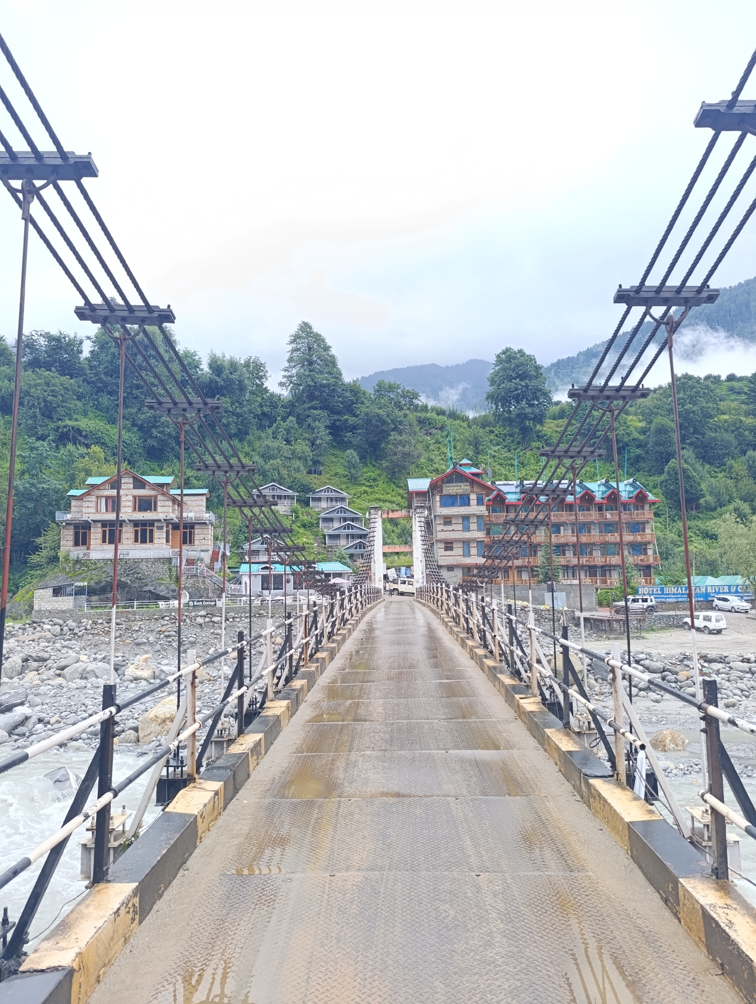 A suspension bridge spans a rocky riverbed under an overcast sky, leading towards a hillside village. The bridge deck is made of diamond-plate metal, wet with visible puddles reflecting the sky. Metal railings with black and white poles line the bridge. On the left bank, a multi-story stone building with a green roof sits atop a rocky slope, with smaller, sloped-roof structures further up the hill. On the right bank, a larger, multi-story hotel with a bright blue roof, featuring numerous windows and balconies, is visible. Signs on this side read "HOTEL HIMALAYAN RIVER & CAFE". Several white vehicles, including a van and cars, are parked on the right side of the bridge, near the hotel. The surrounding environment is characterized by steep, tree-covered hills and a fast-flowing, rocky river. The village appears to be located in Kalath, India, given the presence of buildings and natural scenery typical of the region.
