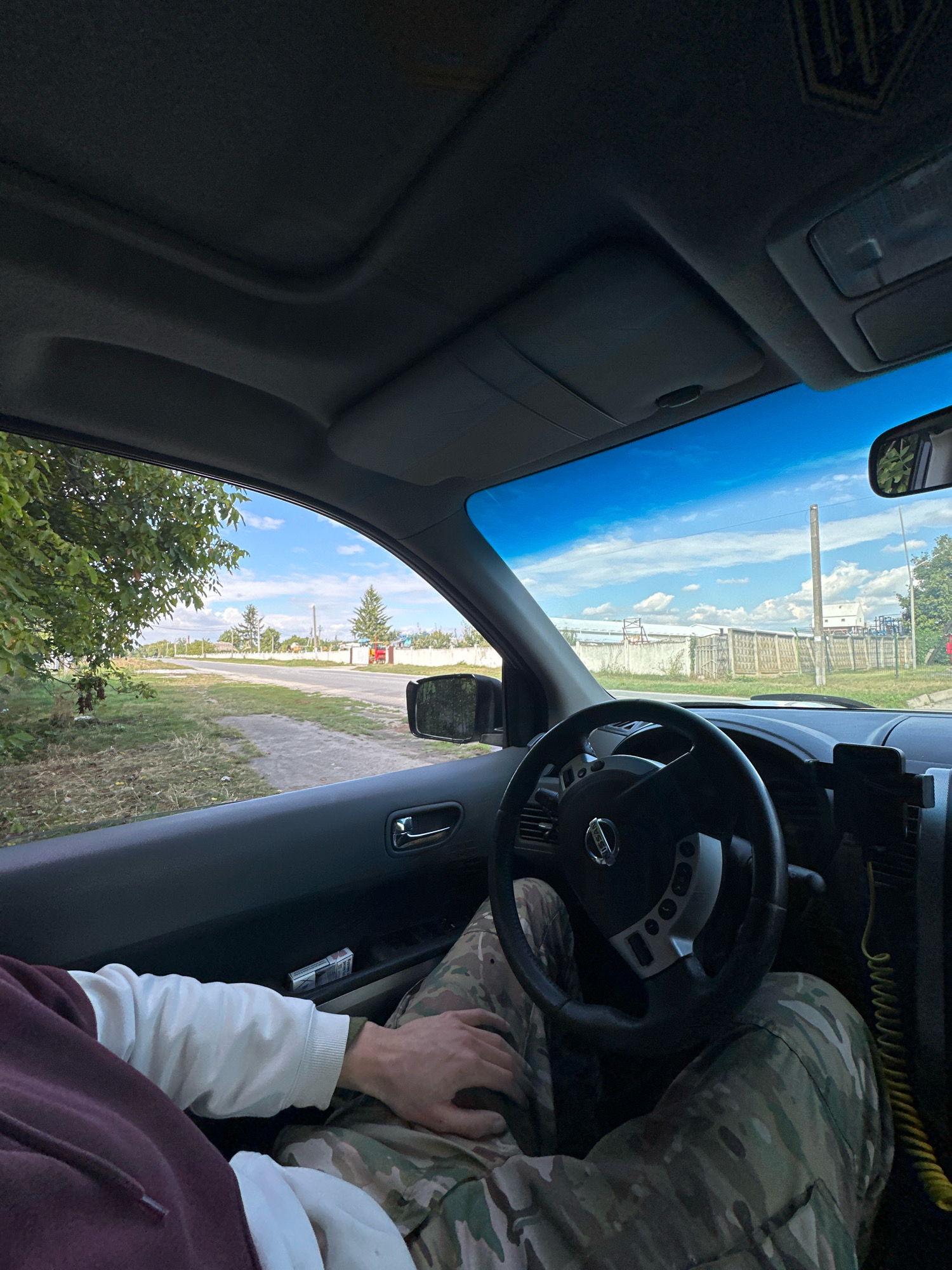 The image shows the interior of a car, looking out the windshield and driver's side window. A person is visible from the waist down, wearing camouflage pants and a maroon hoodie with a white sleeve. Their hands are resting on their lap and the steering wheel. The steering wheel is black and has a Nissan logo. Outside the car, a rural road is visible, lined with trees and houses. The sky is blue with scattered clouds. The location is identified as Shatava, Ukraine.