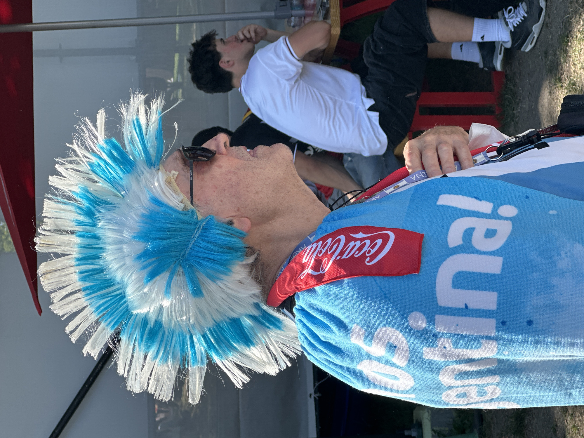 A man with a blue and white mohawk wig, sunglasses, and a blue towel with "Argentina!" printed on it draped over his shoulders stands outside. He is wearing a lanyard with a Coca-Cola logo. In the background, another person in a white shirt sits at a table, and other individuals are visible in the distance. The setting appears to be an outdoor event or gathering. The location is identified as Vancouver, Canada.
