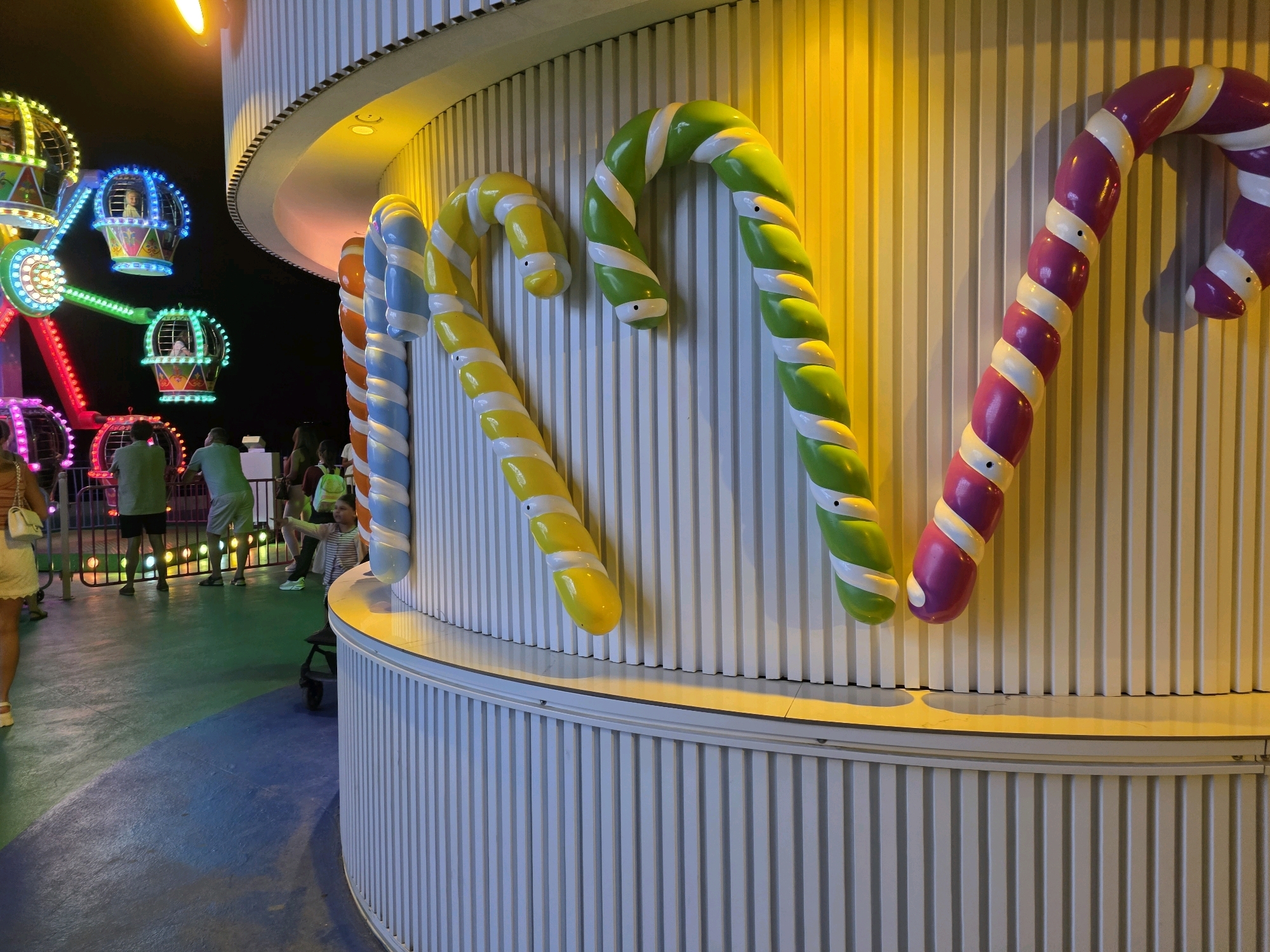 The image depicts a festive scene at an amusement park in Bodrum, Turkey, at night. In the foreground, large, colorful, decorative candy canes are mounted on a white, vertically ribbed surface. From left to right, the visible candy canes are orange and white, blue and white, yellow and white, green and white, and purple and white. The candy canes appear to be made of a glossy material and are illuminated by warm yellow lighting coming from above and behind.
In the background, to the left, a brightly lit Ferris wheel with colorful gondolas can be seen against the dark night sky. Some of the gondolas have children inside. The ground is a multi-colored surface, appearing to be dark blue and green, with some areas illuminated by colored lights from the amusement rides. Several people are visible in the mid-ground, walking and standing, enjoying the park. One person is wearing a white top and yellow shorts, another is wearing a green t-shirt and shorts, and another is wearing a grey t-shirt and shorts. There are also people in the background near the Ferris wheel. The overall atmosphere suggests a lively and cheerful nighttime amusement park experience.