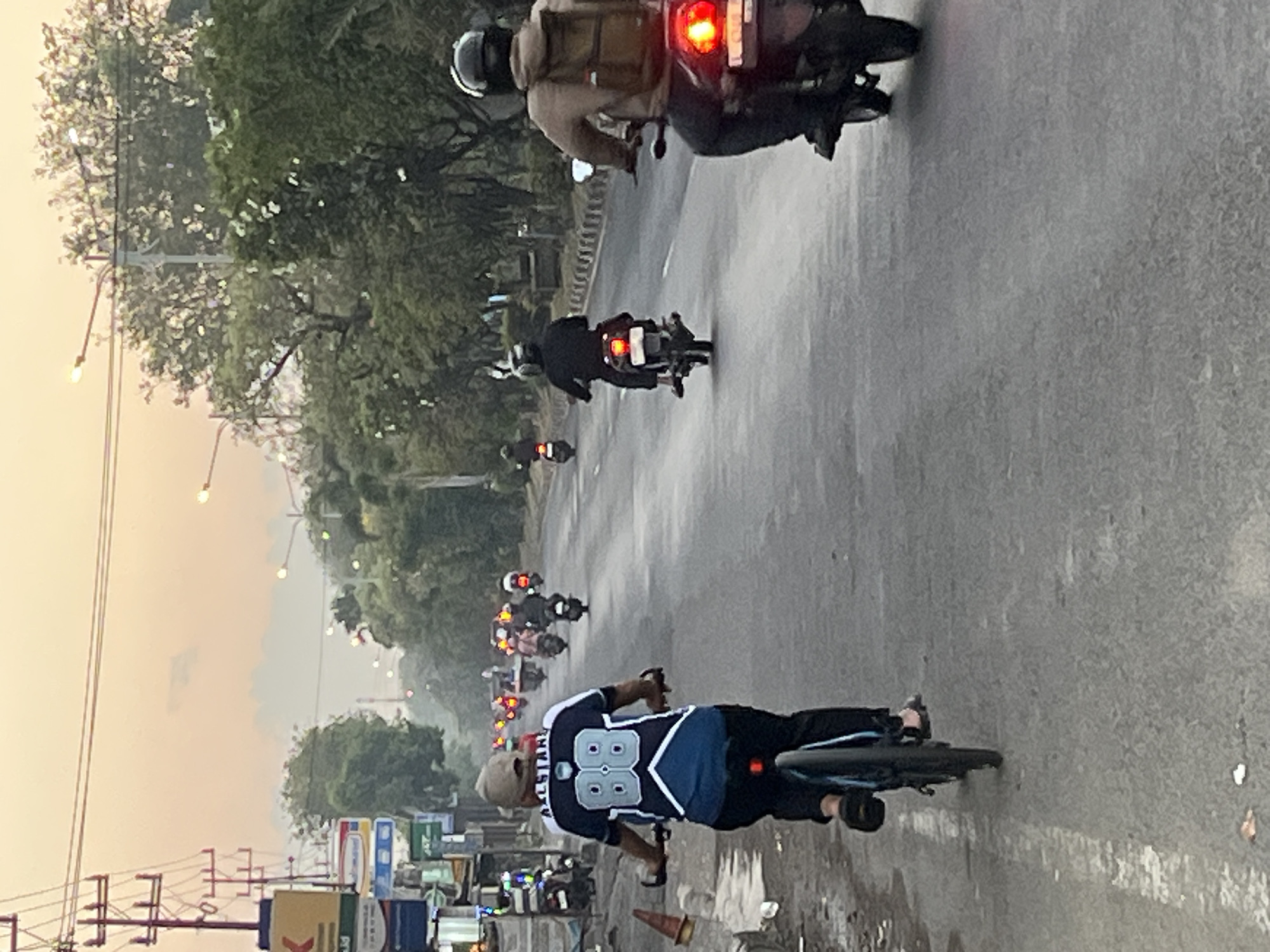 In Ngaban, Indonesia, on a paved road with a white line marking, a group of people are riding motorcycles and a bicycle. The scene is set against a dusky sky with power lines and trees lining the sides of the road.
In the foreground, a person is riding a bicycle. They are wearing a light-colored cap and a dark blue jersey with white accents and the number "88" prominently displayed on the back. They are also wearing dark pants and sandals. The bicycle has a blue frame and black tires.
Further down the road, several individuals are riding motorcycles. One motorcyclist in the middle distance wears a black helmet and a dark jacket. Another motorcyclist, closer to the foreground on the right, is wearing a brown jacket and a backpack. Their motorcycle has a red taillight. In the background, more motorcycles are visible, their taillights illuminated.
On the left side of the road, there are buildings with signs and some parked motorcycles. The environment appears to be a somewhat busy urban or suburban area. The overall lighting suggests it is either early morning or late afternoon.