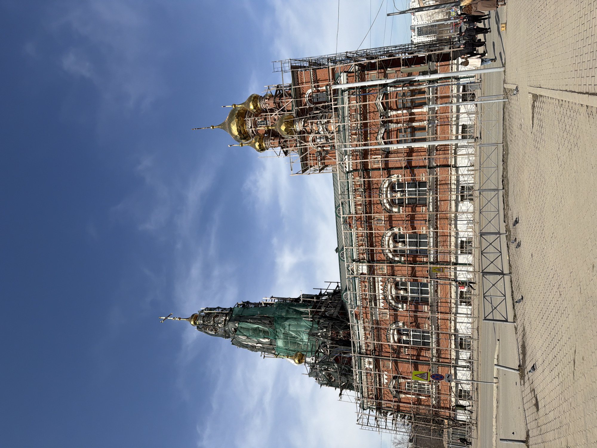 The image depicts a brick building in Perm, Russia, under construction or renovation. Scaffolding covers much of the building's facade. The building features prominent golden domes, partially covered with green tarp on one side. The sky is visible with scattered clouds. A sidewalk and street are present. A group of people are visible on the sidewalk. Metal barriers are positioned along the sidewalk's edge. A road sign is visible near the building. The orientation of the image is vertical.