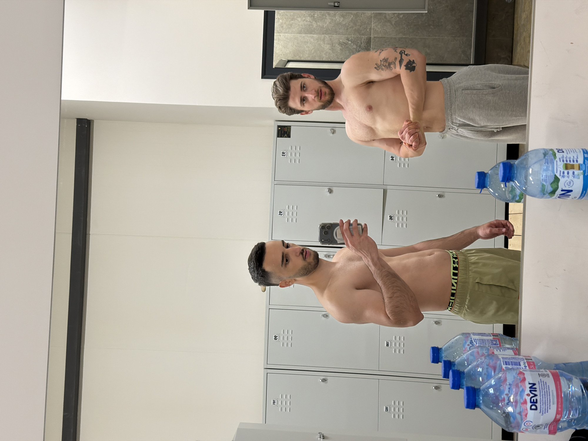 The image, rotated 90 degrees clockwise, depicts two shirtless men captured in a mirror reflection within a clean, modern locker room. The setting is characterized by rows of light grey metal lockers with visible numbers, including "83" to "88" and "100" to "105". A bright white ceiling and darker tiled walls are also reflected.
In the foreground of the reflection, a man with dark hair and a beard, wearing olive green shorts with an "UNDER ARMOUR" waistband, holds a smartphone, seemingly taking a mirror selfie. Further into the reflection, another man with lighter hair and a beard, wearing grey sweatpants, is posing, flexing his biceps and looking towards the camera. He displays several tattoos on his left arm, including floral designs and a stylized dagger.
Several plastic water bottles are lined up on a reflective surface in the foreground. The labels on these bottles clearly show the brand "DEVIN" and text indicating "1900 ml" in Cyrillic. The scene suggests a post-workout or pre-workout moment. This locker room is located in Plovdiv, Bulgaria.