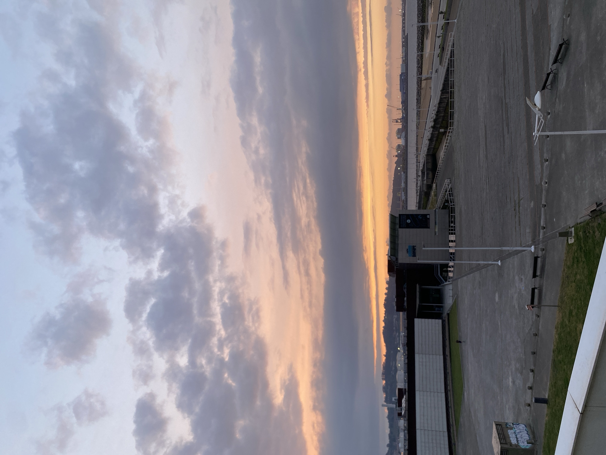 The image captures a wide panoramic view of a coastal urban landscape during either dawn or dusk, indicated by the warm, vibrant colors of the sky. The horizon line runs horizontally across the middle of the frame, dividing the expansive sky from the ground and urban structures.
The sky is a dominant feature, displaying a beautiful gradient of colors. In the upper left, it is a soft blue with scattered, fluffy grey and white clouds. Towards the horizon, these colors transition into striking oranges, yellows, and soft pinks, creating a luminous glow that suggests the presence of the sun just below the horizon. Darker, more elongated clouds stretch across the warmer parts of the sky, adding texture.
Below the sky, the scene unfolds with paved areas, structures, and distant features. In the immediate foreground (bottom left after rotation), there's a paved walkway or plaza, possibly concrete, with a white railing or wall. To its right, the paved area continues, flanked by patches of green grass. Several modern lamp posts with white fixtures and dark benches are visible on the paved ground.
Further into the mid-ground, a large, dark building with glass elements is situated, possibly a public building or cultural center. On its facade, indistinct white text is visible, including "LA RUTA" and "FRONTERA" (or similar), and "LA CASA". Another building with a dark facade and white signage, possibly reading "MUSEO" or "MOORE", is also present. On a low concrete structure, some illegible graffiti is visible.
Along the horizon, a body of water, likely the sea or an estuary, reflects the sky's colors faintly. On the far side of the water, a distant landmass or shoreline is silhouetted, dotted with artificial lights and faint outlines of industrial structures, including cranes, suggesting a port or harbor. One or two small, distant figures can be seen on the paved ground, but they are not central to the scene.
The overall atmosphere is serene and contemplative, highlighting the beauty of the twilight sky over a modern coastal environment, consistent with the location of Gijón/Xixón, Spain.