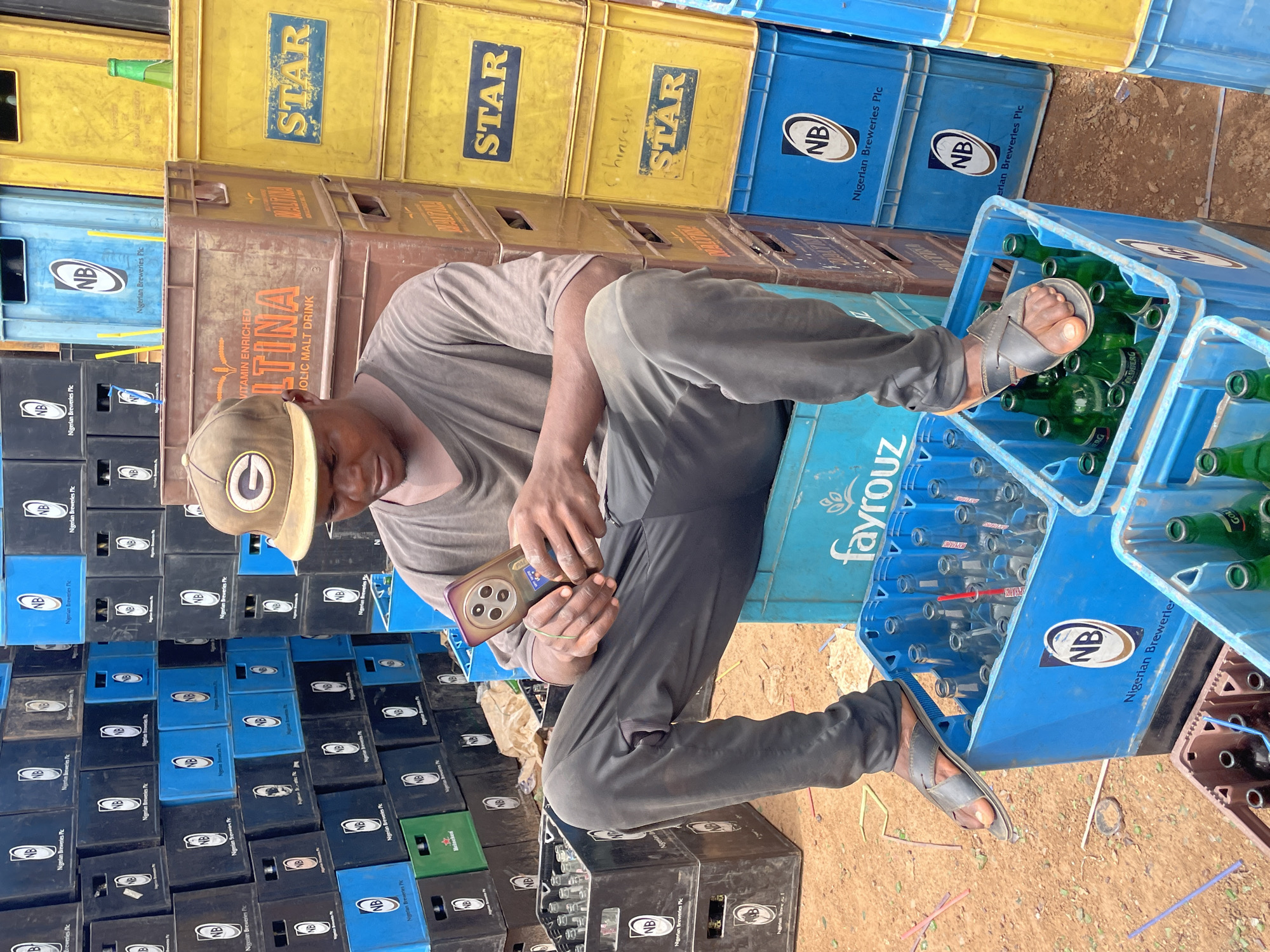 A man wearing a tan t-shirt, gray pants, and a tan baseball cap with a green emblem sits on a blue plastic beer crate. He is holding a smartphone in his hands. The man is surrounded by stacks of plastic beer crates in various colors, including yellow, brown, blue, and black. Some of the crates contain empty green beer bottles. The crates are labeled with various brand names, including "STAR," "LTINA," "fayrouz," and "NB Nigerian Breweries Plc." The scene appears to be located in Bukuru, Nigeria.