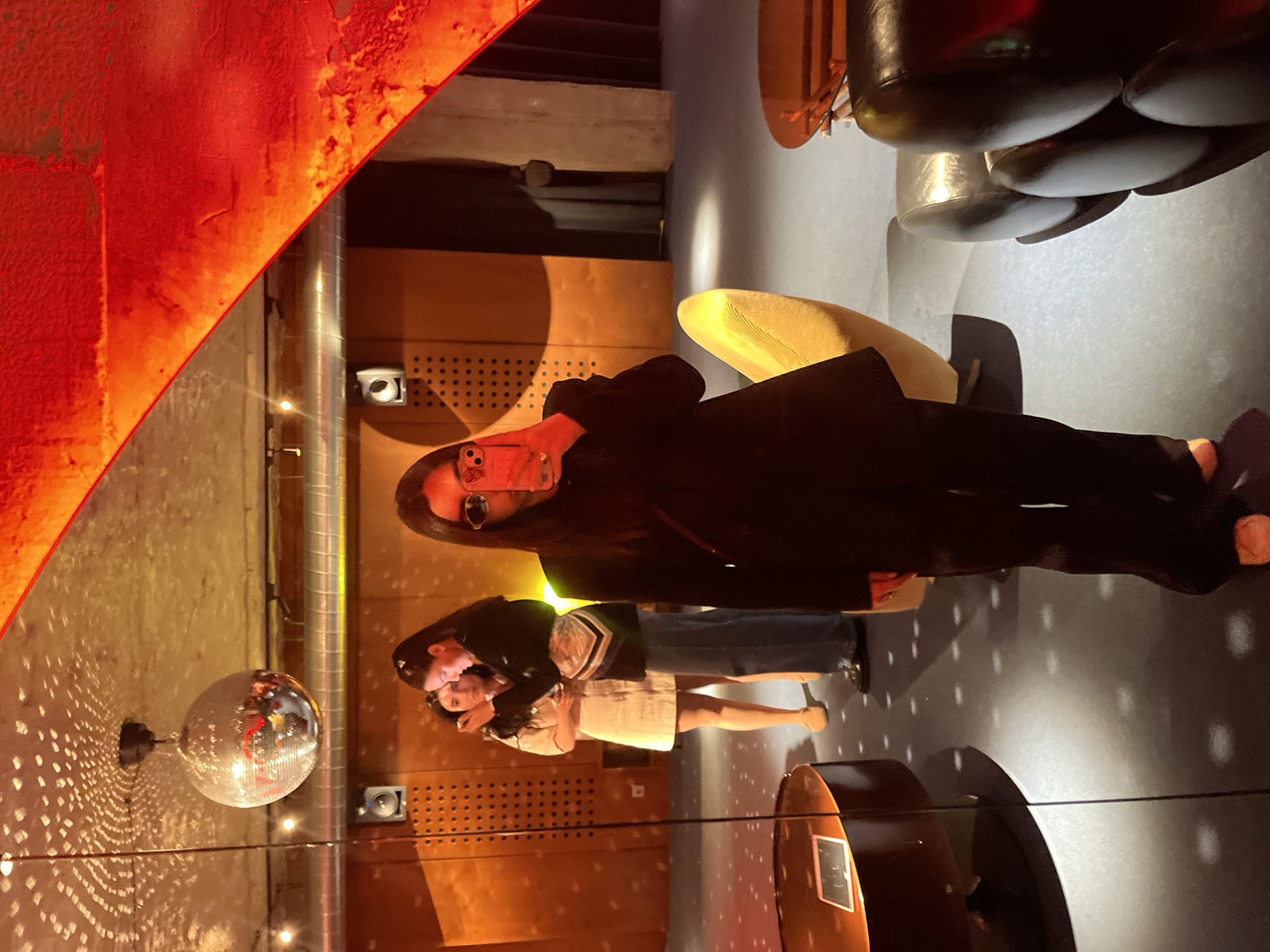 Two women pose in a room illuminated by a disco ball in Yerevan, Armenia. One woman, wearing glasses and a black jacket, holds a phone to her face. The other woman embraces her and wears a white dress and dark jacket. The room features a red, curved structure, a yellow chair, and lighting fixtures on the wall.