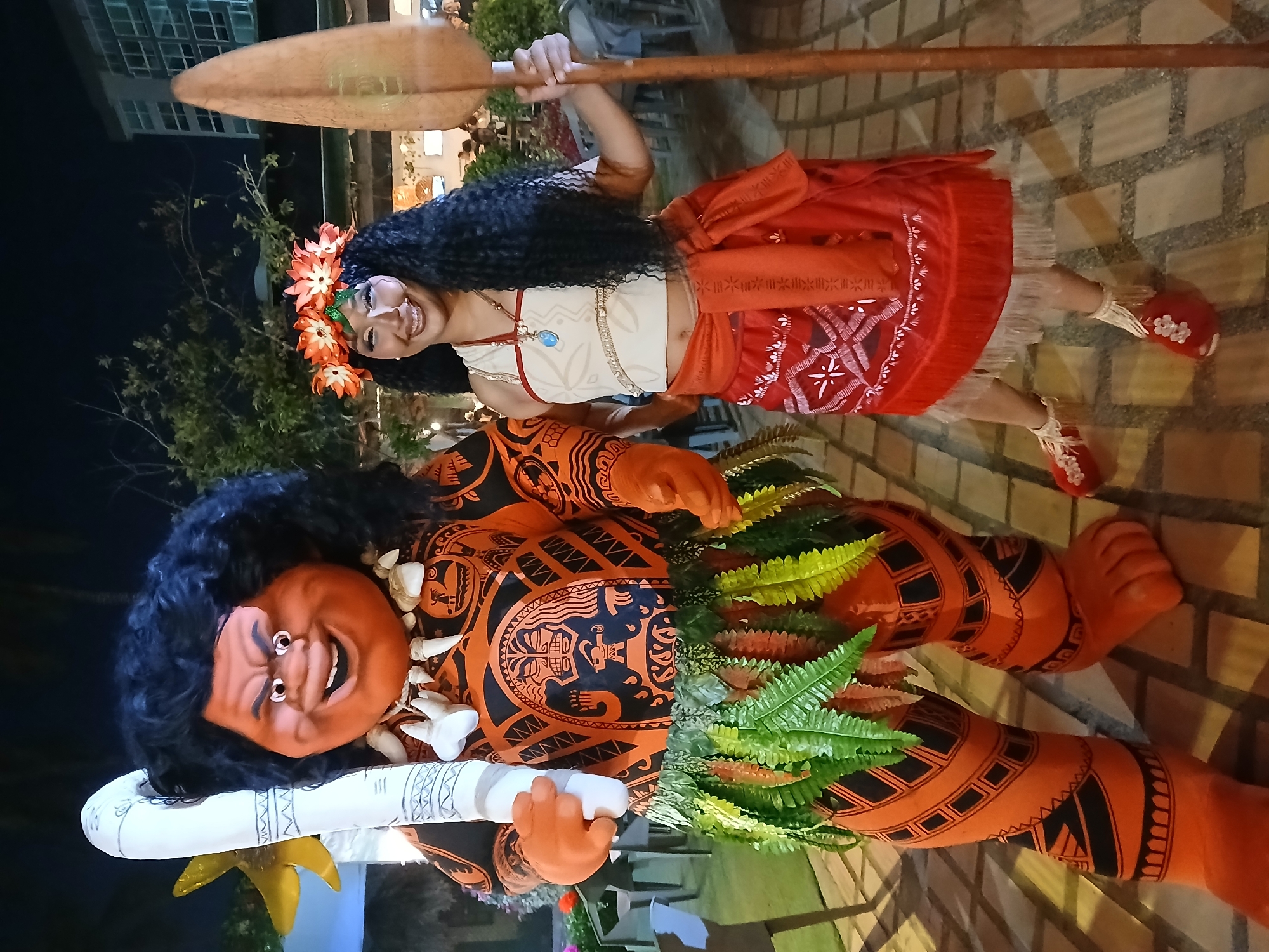 A woman dressed as Moana holds a paddle behind her back, standing beside a figure resembling Maui from the Disney film "Moana." The Maui figure is lying down, clad in orange and black patterned clothing, with a green fern skirt and holding a white club. Both are positioned on a paved area, likely in Porlamar, Venezuela.