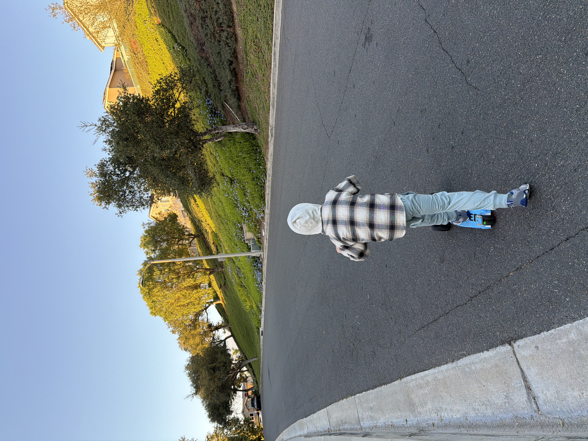 A young child wearing a plaid shirt and light blue pants is riding a blue scooter on an asphalt street in Aliso Viejo, California. The street curves uphill and is lined with a concrete curb and a grassy slope with trees and houses on top. The sky is clear and blue, and the sun is casting long shadows.