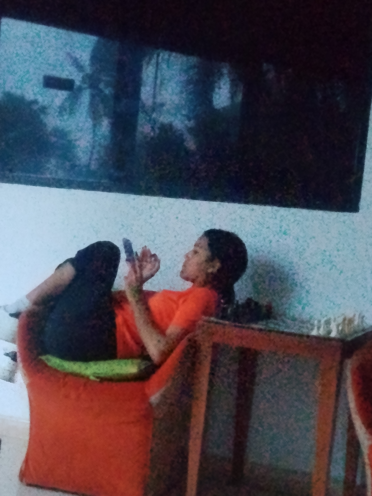 A person wearing an orange shirt and black pants is sitting on an orange chair, looking at their phone. The person is in a room with a television screen on the wall showing a jungle scene. There is a wooden table in the foreground. The room appears to be located in Playa El Agua, Venezuela.