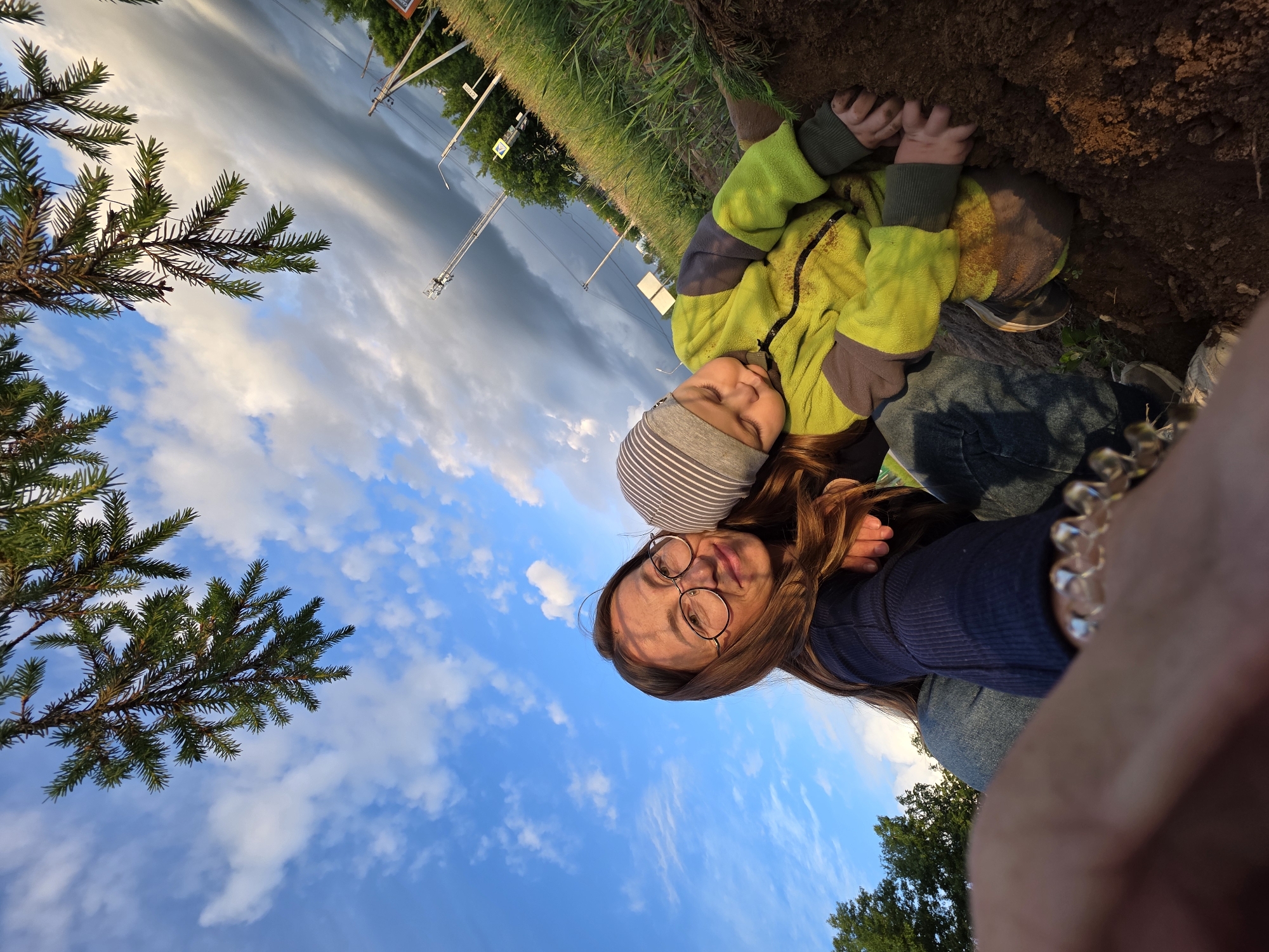 The image shows a woman and a child in an outdoor environment. The woman is wearing glasses and a blue shirt. The child is wearing a striped hat and a yellow jacket. They appear to be near a patch of dirt or soil and some vegetation. In the background, there is a cloudy sky and what appears to be roadside greenery and a structure with poles. Considering the prompt request of the location as Chaikovsky, Russia, the roadside structure with poles could suggest street lighting or utility infrastructure common in urban or suburban areas of the city. Based on the angle and composition, it seems like a selfie-style photo.