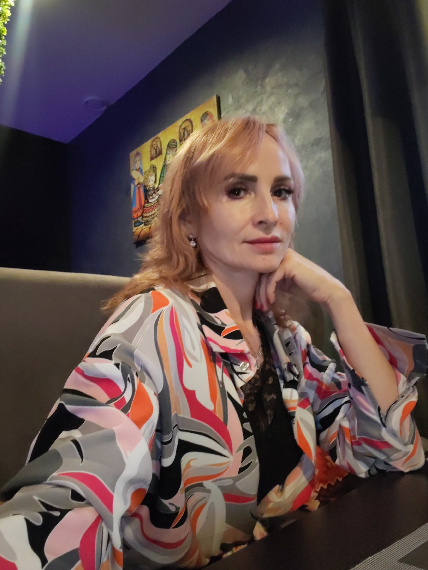 A woman with reddish-blonde hair, wearing a colorful patterned blouse over a black lace top, poses for a selfie in what appears to be a restaurant. She has her right hand resting on her chin and a faint smile on her face. Behind her, a painting of people in traditional attire hangs on a dark blue wall. A purple light illuminates the upper left corner of the frame, and dark curtains are visible to the right. The setting suggests it might be evening, as indicated by the artificial lighting. There are no visible text elements in the image.