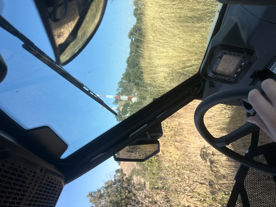 The view is from inside a utility terrain vehicle (UTV). A windshield wiper is visible against a clear blue sky. A red and white tower stands in the distance. The terrain outside is dry, grassy, and hilly. A side mirror is mounted on the vehicle's frame. The driver's hand rests on the steering wheel, with a digital display visible on the dashboard. The image suggests the vehicle is traversing an arid, outdoor environment.