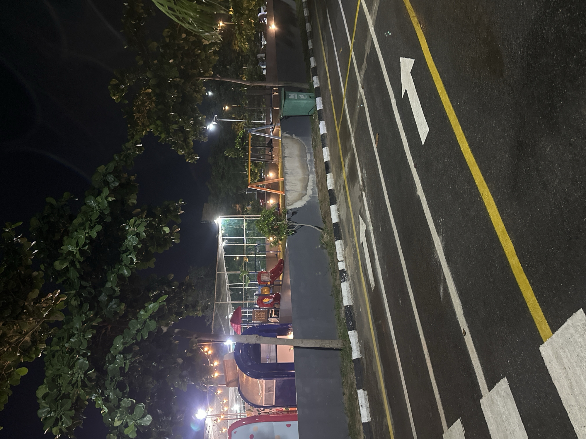 The image depicts a nighttime scene at a playground in Abuja, Nigeria. A dark asphalt road with white and yellow lane markings, including a directional arrow, dominates the foreground. Beyond the road, a low gray wall separates it from a playground area. The playground features a blue tunnel slide, red and yellow playground equipment, and a swing set with an orange frame. Trees with lush green foliage are scattered throughout the scene, and string lights illuminate the playground area. A green trash bin is visible near the wall. The overall environment suggests an outdoor recreational space illuminated for evening use. There are no people visible in the image.