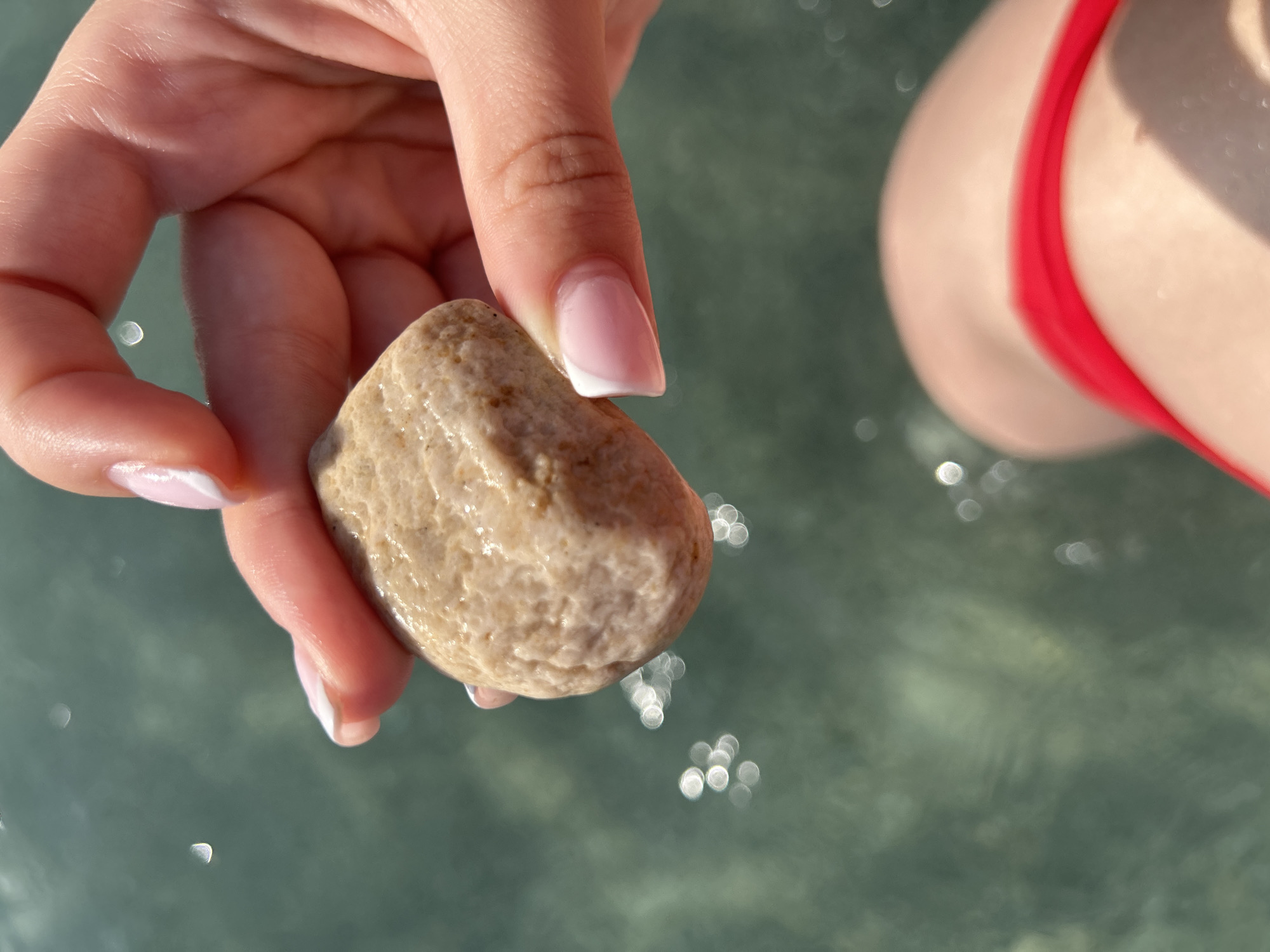 A hand with manicured nails is holding a smooth, beige-colored rock. The background shows blurry water with bokeh effects and a glimpse of a red bikini top. The location is not specified within the city of Unknown, Unknown.