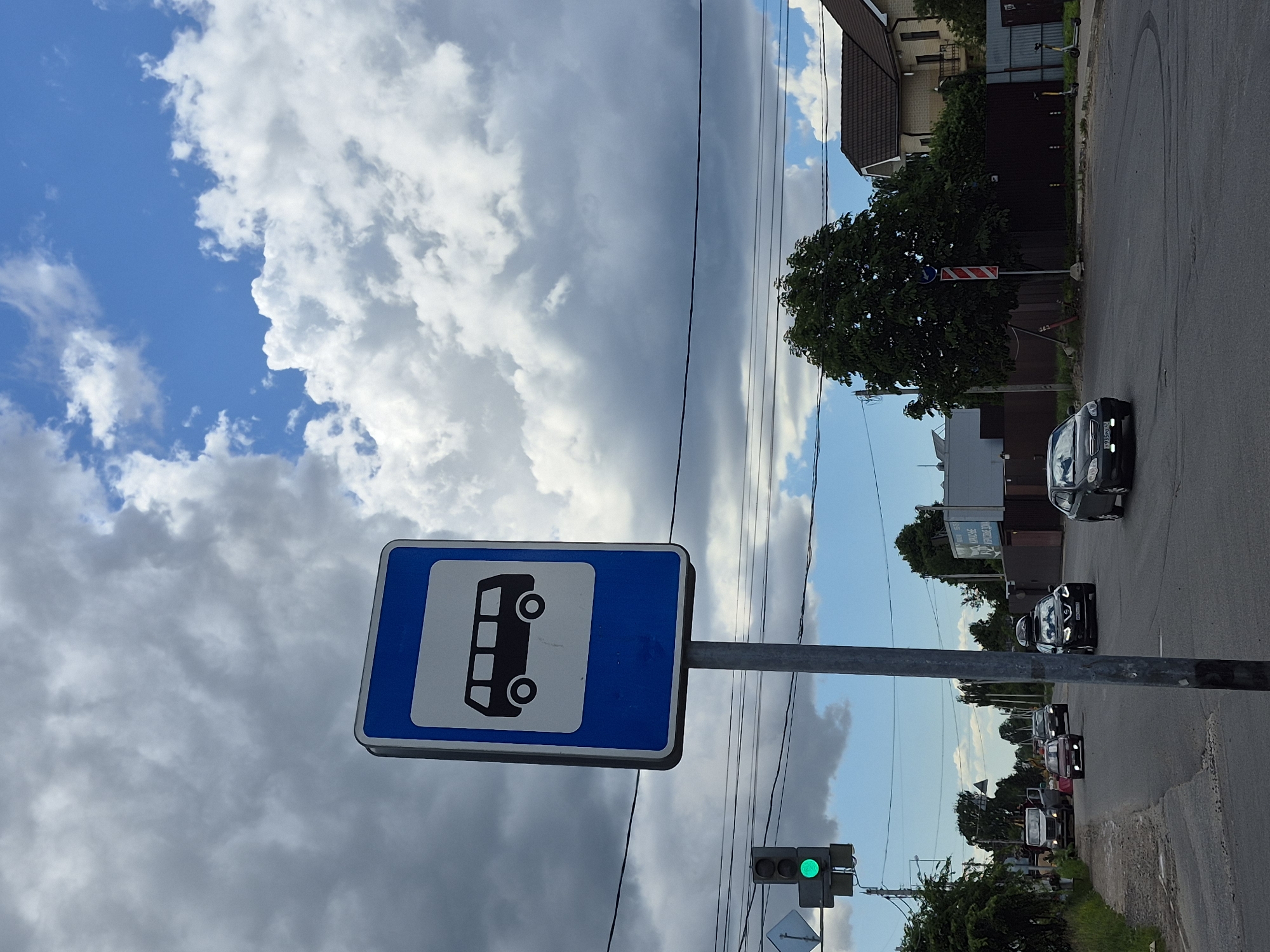 The image depicts a street scene in Koltushi, Russia, featuring a bus stop sign with a pictogram of a bus. The sign is mounted on a metal pole. To the left of the bus stop sign, a traffic light displays a green signal. Several cars are visible on the road, including a black SUV with a roof rack, a dark-colored sedan, and a red sedan. In the background, there are buildings with residential architecture, trees, and a cloudy sky. The road appears to be an intersection. The scene is set outdoors under a partly cloudy sky.