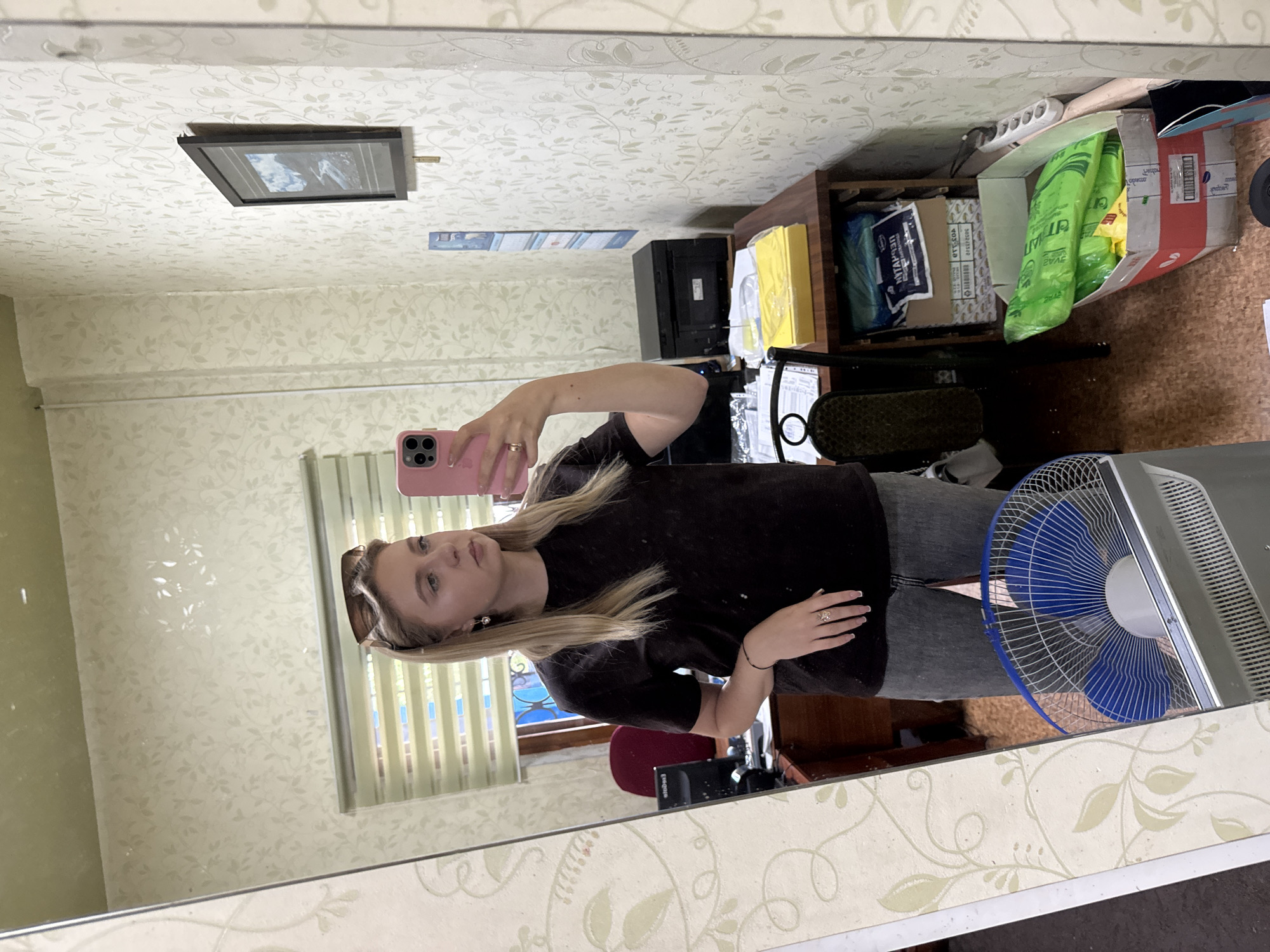 A young blonde woman is taking a mirror selfie in a room that appears to be an office or study. She is wearing a black t-shirt and grey jeans, and has sunglasses perched on her head. In her right hand, she holds a pink smartphone. The room's walls are covered in a light-colored, floral-patterned wallpaper. To the left of the woman, a desk is visible with a computer monitor and a red chair. To her right, a shelf unit holds various office supplies and boxes, including a black printer and a box of green plastic bags. A blue and white electric fan is positioned in the foreground, also reflected in the mirror. The overall scene suggests a casual, everyday moment captured in a somewhat cluttered domestic or work environment.