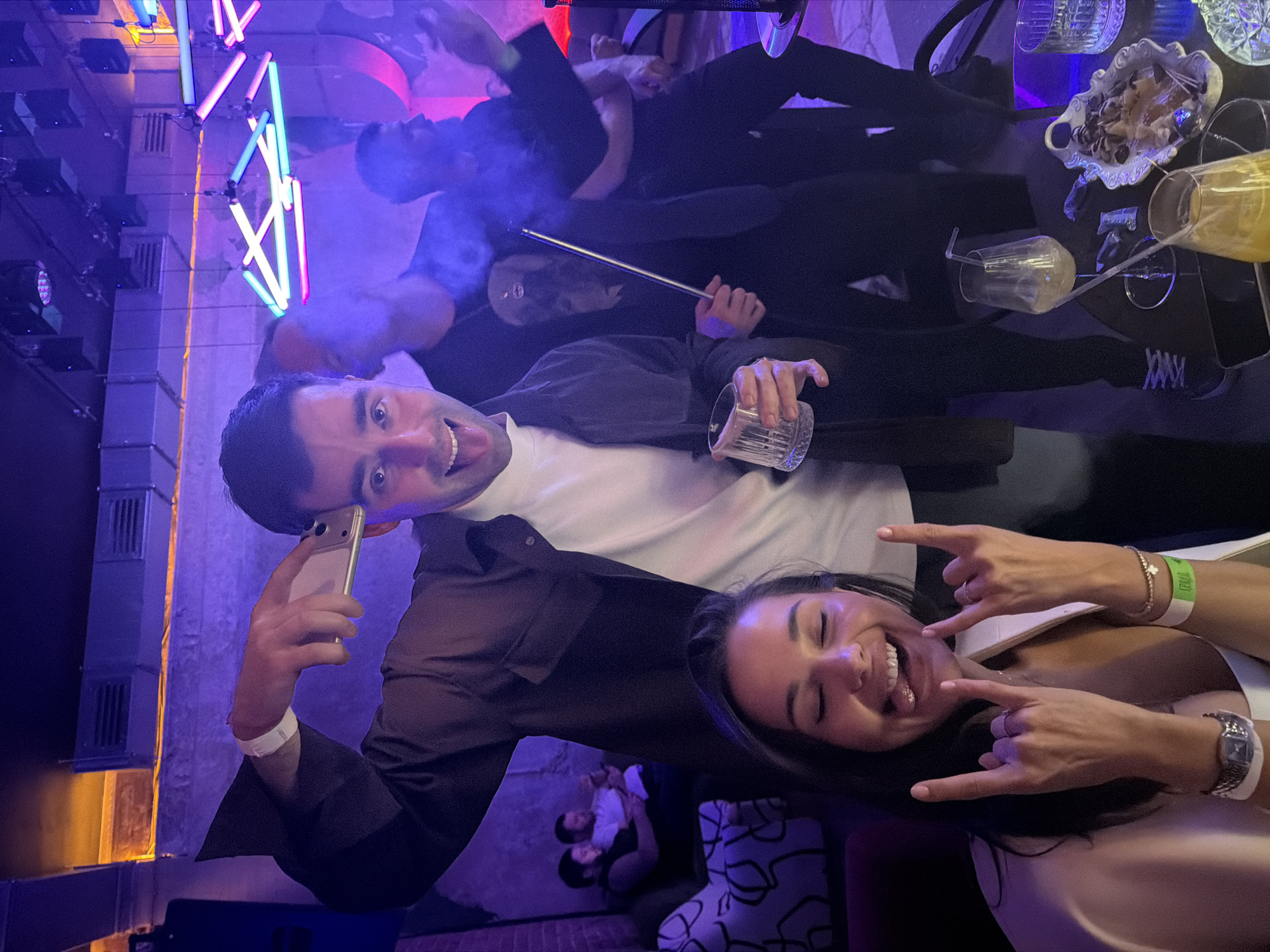The image captures a vibrant night scene inside a lively bar or club in Krasnoyarsk, Russia. The setting is dimly lit with colorful artificial lighting, including decorative neon tube lights in shades of blue, pink, purple, and green, creating an energetic and modern ambiance.
In the foreground, a man and a woman are posing for the camera, both expressing high spirits. The man, with dark hair, smiles widely with his tongue slightly out, holding a smartphone in his left hand and a glass containing a drink in his right. He wears a dark shirt over a white T-shirt and a white wristband. Beside him, a woman with dark hair smiles joyfully with her eyes closed, making "rock on" hand gestures with both hands. She wears a light-colored top, a watch on her left wrist, and a green wristband that clearly displays the text "I ❤ BARS".
Several other people are visible in the midground and background, indicating a social gathering. One individual is seen smoking a hookah, with a cloud of smoke or vapor around them. Tables are adorned with drinks in various glasses, some holding a yellowish liquid, and a white decorative bowl filled with snacks like nuts or candies. The activity suggests a festive and relaxed social event, typical of nightlife. The image is rotated 90 degrees clockwise, presenting a dynamic, tilted perspective of the bustling indoor environment.
