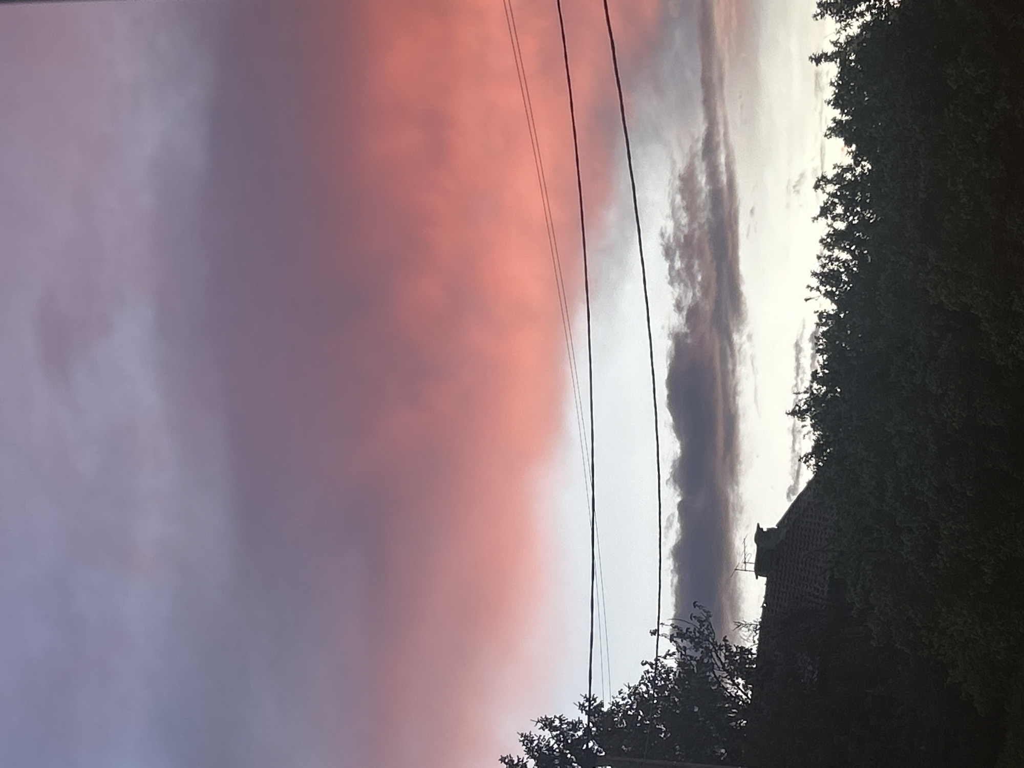 The image depicts a colorful sky at dusk or dawn in the city of Unknown, Unknown. The sky transitions from a deep purple on the left to a pinkish-orange hue, with a narrow band of white and gray clouds in the middle. Power lines run diagonally across the center of the frame. Trees and part of a building are silhouetted against the sky on the right side. The building appears to be a house with a roof.